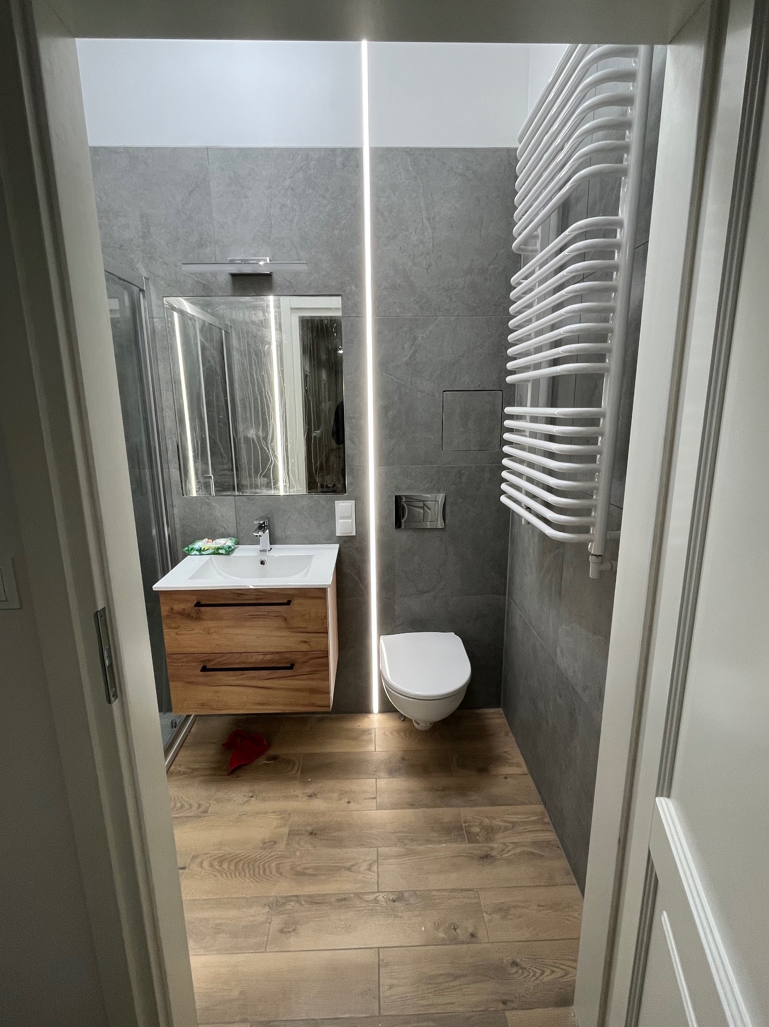 Nowoczesna łazienka z szarymi płytkami, drewnianą szafką pod umywalką i grzejnikiem drabinkowym. Uwagę przyciąga pionowe oświetlenie LED wzdłuż ściany.