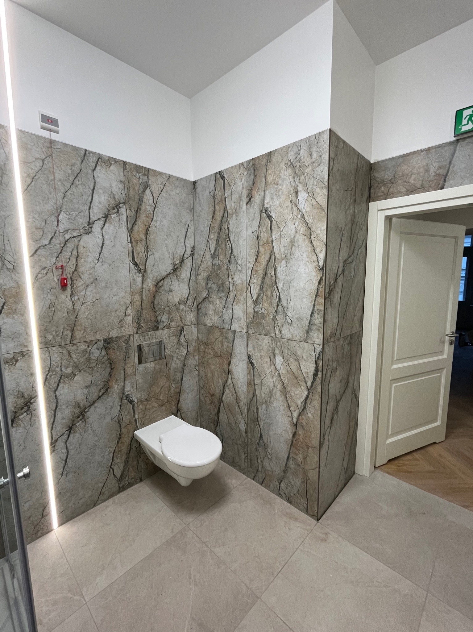 Nowoczesna łazienka z marmurowymi płytkami ściennymi, podwieszaną toaletą i subtelnym oświetleniem LED. Widoczny fragment kabiny prysznicowej i otwarte drzwi.