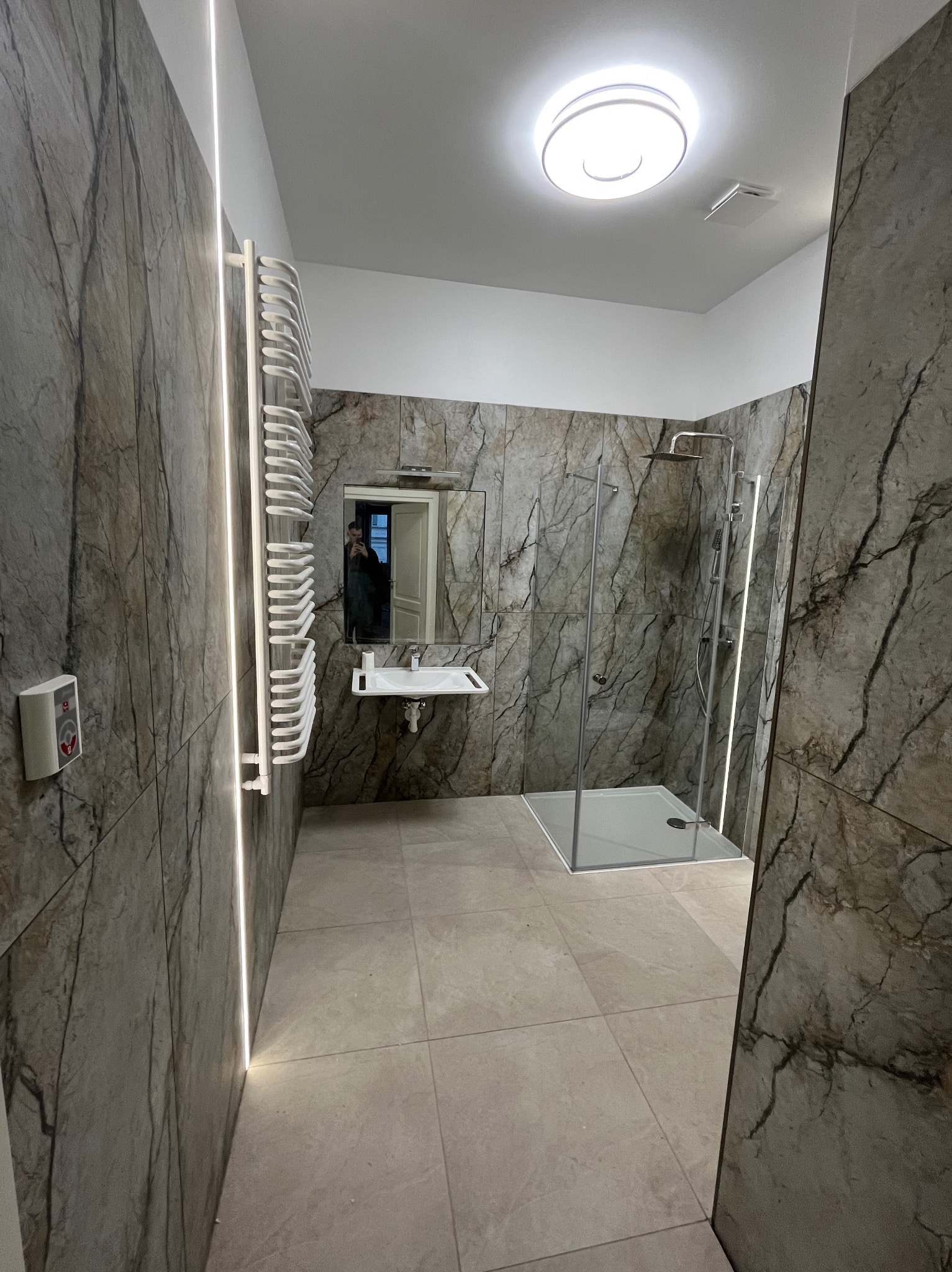 Wykończona łazienka z kabiną prysznicową, umywalką i grzejnikiem drabinkowym. Ściany wyłożone płytkami imitującymi marmur, oświetlenie LED wzdłuż krawędzi płytek.