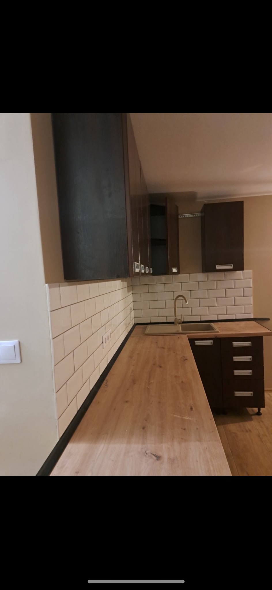 Nowoczesna kuchnia z drewnianym blatem i białymi kaflami. Ciemne szafki wiszące i stojące. Zlewozmywak ze złotą baterią. Widoczne gniazdka elektryczne.