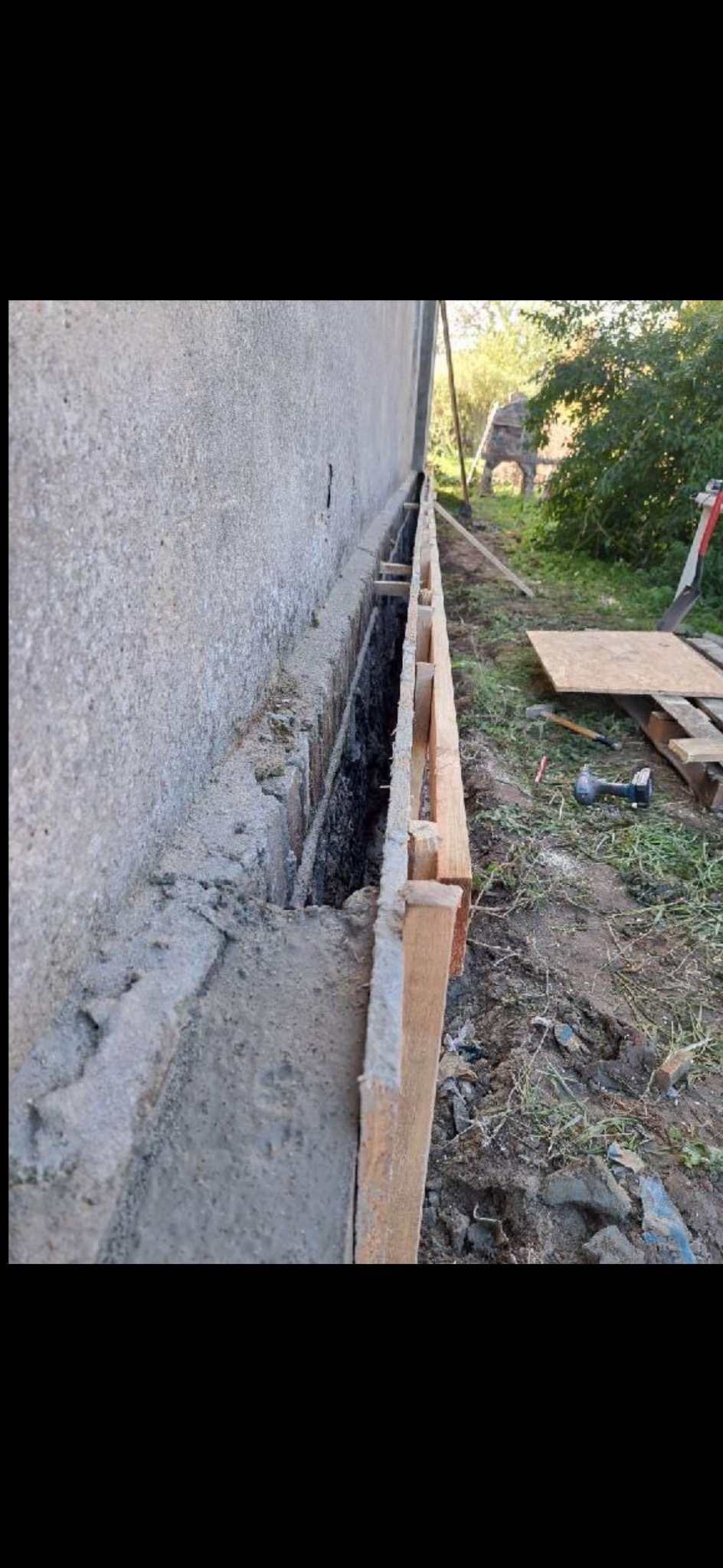 Fundament budynku w trakcie wzmacniania, z widocznym szalunkiem i świeżym betonem. Na zewnątrz narzędzia budowlane i zielona roślinność.