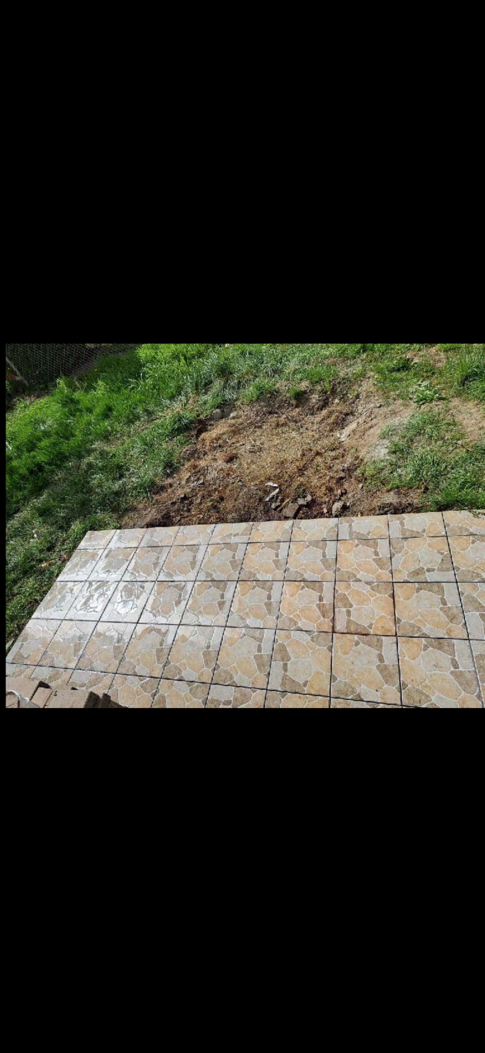 Ułożona, beżowa, kwadratowa terakota na zewnątrz, przylegająca do trawnika. Widok z góry, na pierwszym planie płytki, w tle trawa i ziemia.