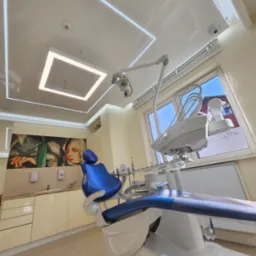 Nowoczesny gabinet dentystyczny z sufitem napinanym i oświetleniem LED, mozaiką na ścianie i fotelem dentystycznym w kolorze niebieskim. Jasne, przestronne wnętrze.