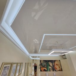 STRETCHART SPÓŁKA Z OGRANICZONĄ ODPOWIEDZIALNOŚCIĄ - Biały, błyszczący sufit napinany z oświetleniem LED w kształcie prostokątów. W tle widoczna osoba robiąca zdjęcie i certyfikaty na ścianie.