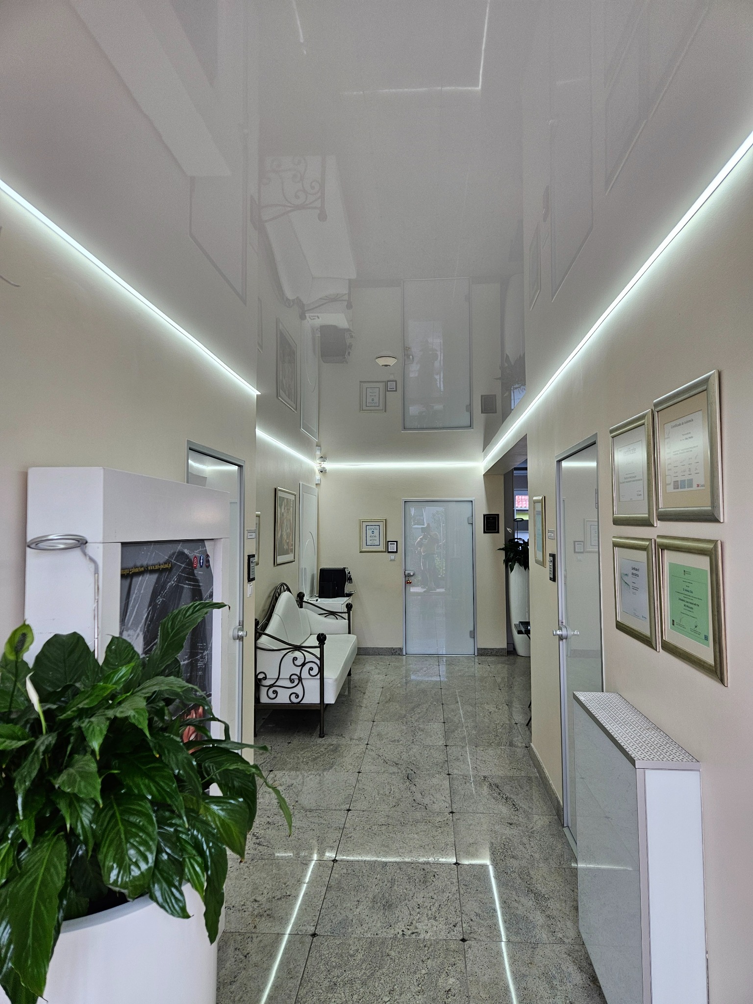 Biały, błyszczący sufit napinany z oświetleniem LED wzdłuż korytarza biura, odbijający wnętrze. Na ścianach certyfikaty i dyplomy, podłoga z szarego kamienia.