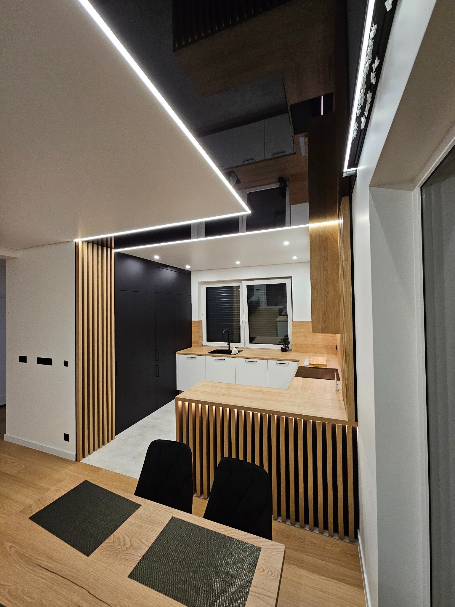 Nowoczesna kuchnia z czarnymi szafkami i drewnianymi lamelami, oświetlenie LED w suficie i nad blatem, widok na stół z krzesłami.