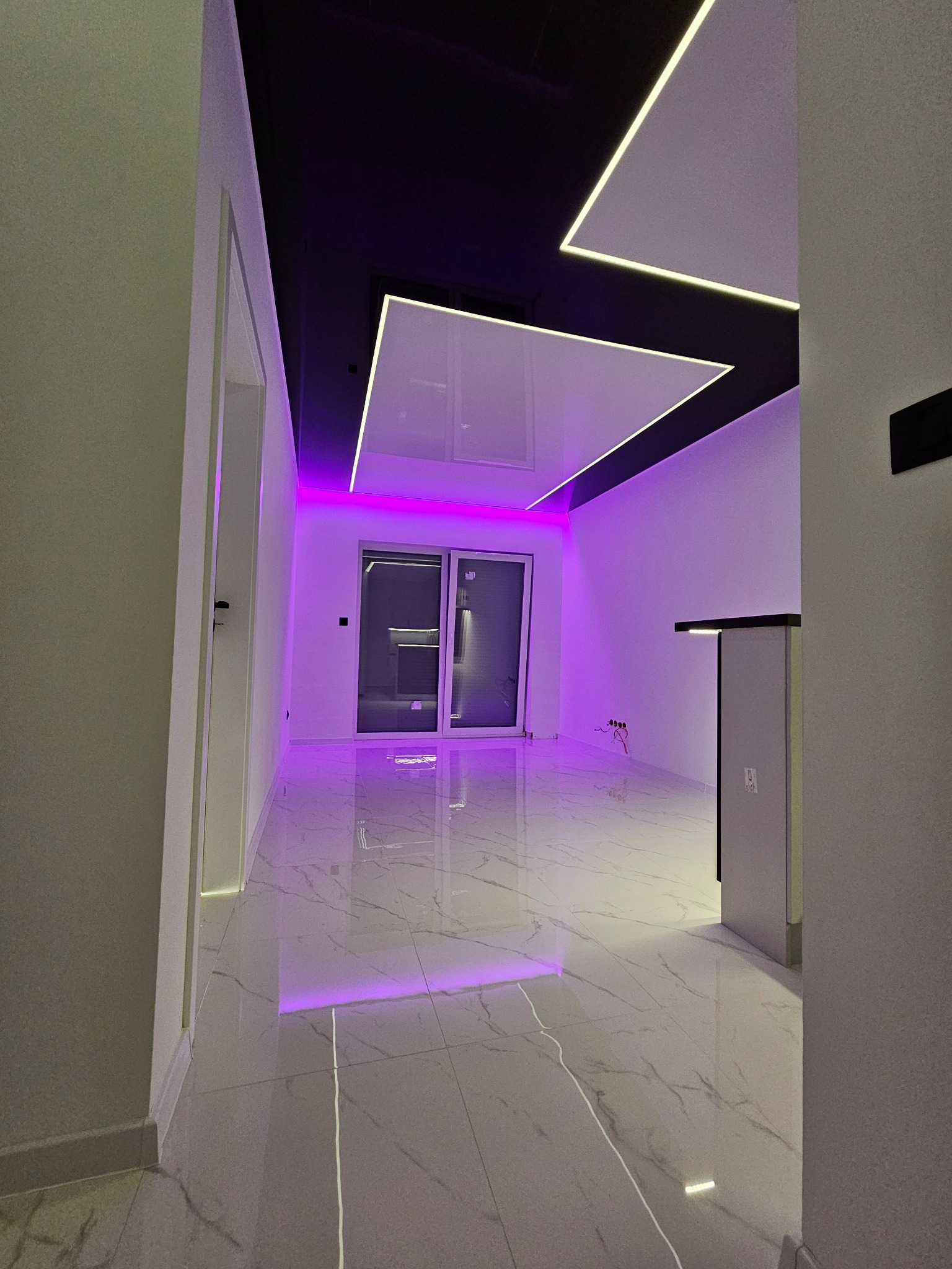 Nowoczesne wnętrze z podświetlanym sufitem napinanym, fioletowe akcenty świetlne, marmurowe płytki podłogowe i minimalistyczny design. Widok na przesuwne drzwi balkonowe.