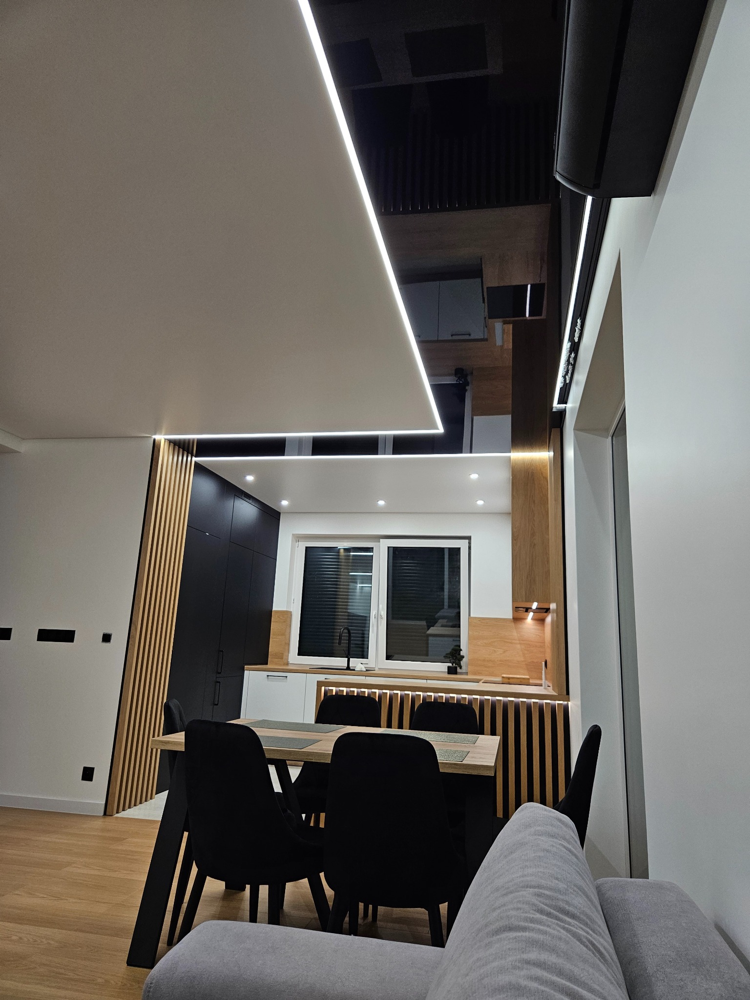 Nowoczesne wnętrze: biały sufit napinany z oświetleniem LED, czarny, lustrzany sufit nad stołem jadalnym, drewniane lamele na ścianie i zabudowa kuchenna.