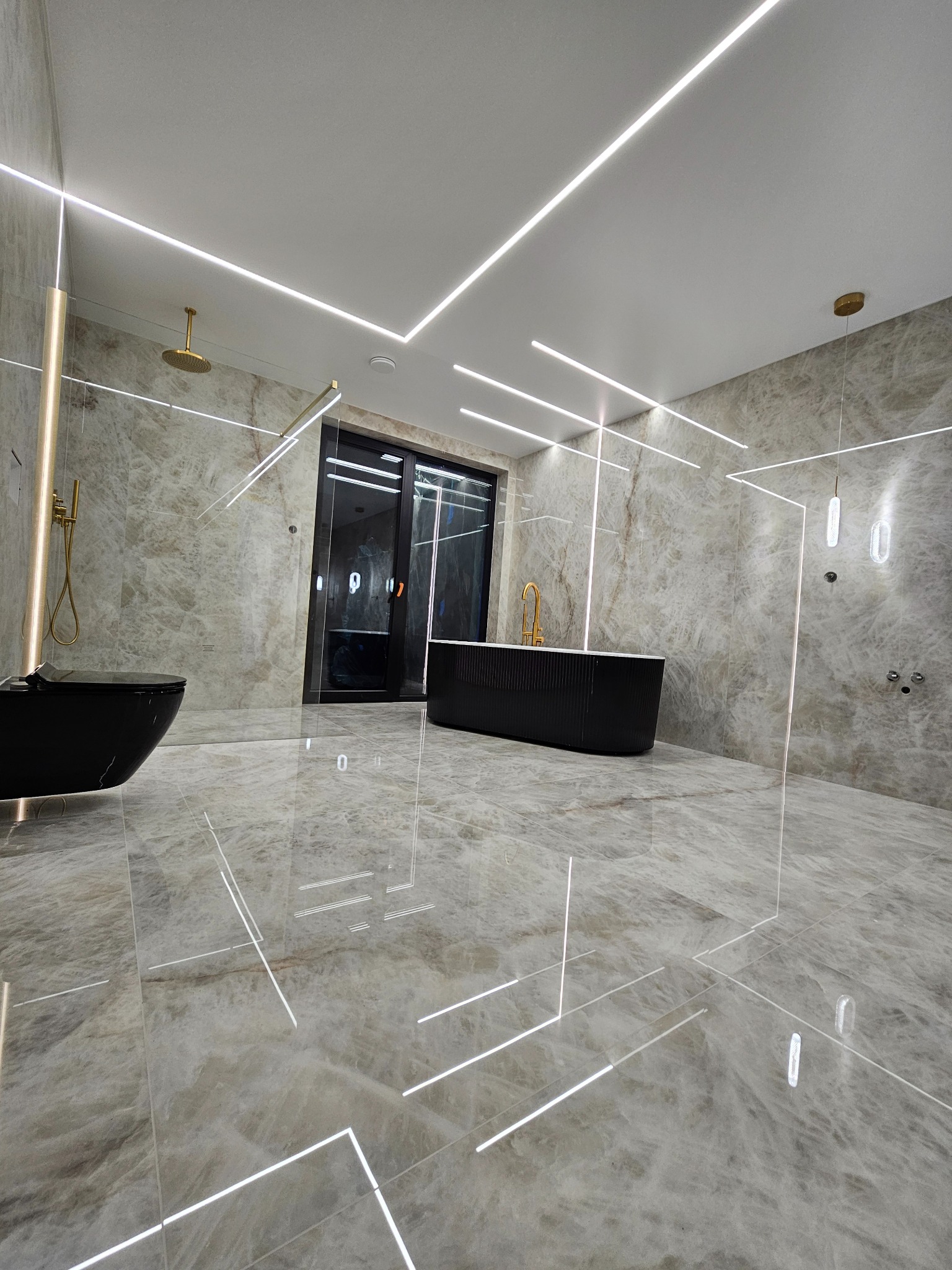 Elegancka łazienka z marmurowymi płytkami, czarną armaturą i liniowym oświetleniem LED. Minimalistyczny design z wanną wolnostojącą i kabiną prysznicową typu walk-in.