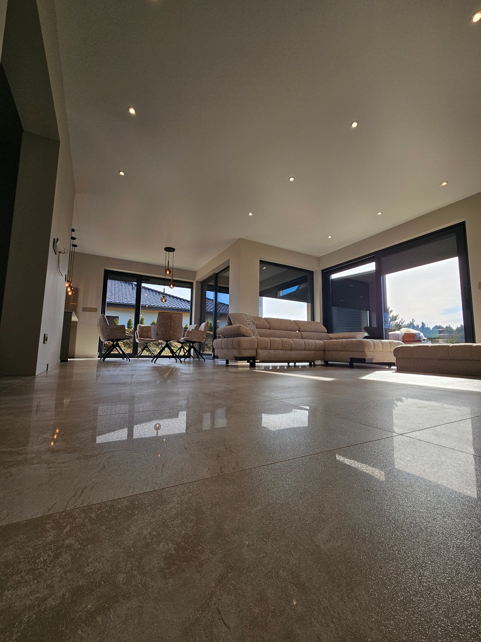 Przestronny salon z beżową sofą, jasnymi płytkami podłogowymi i dużymi oknami. Widoczne designerskie lampy i krzesła. Minimalistyczny wystrój wnętrza.