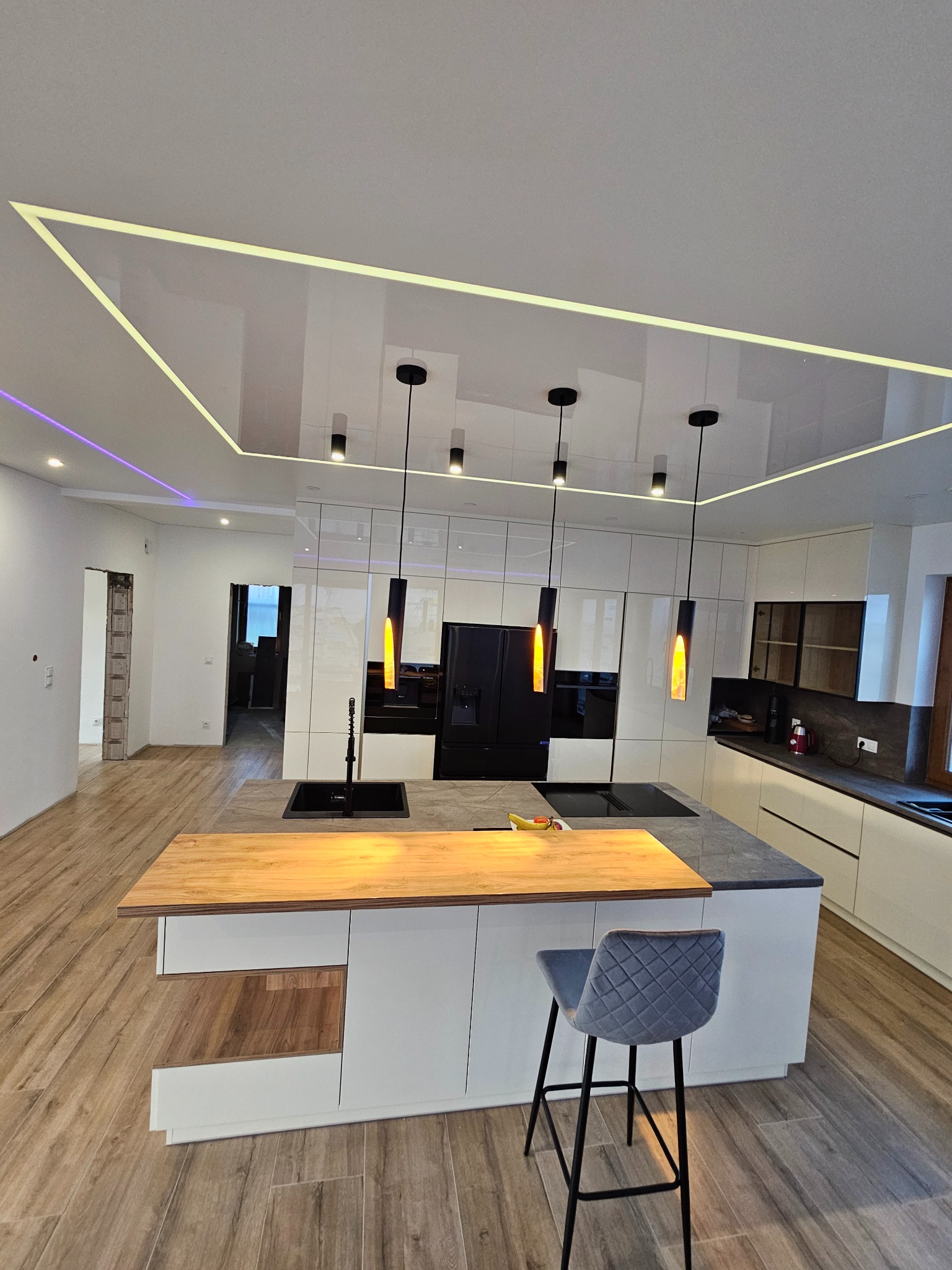 Biała kuchnia z wyspą i szarym krzesłem barowym. Sufit napinany z oświetleniem LED w kształcie prostokąta. Nowoczesny design, jasne kolory, drewniana podłoga.