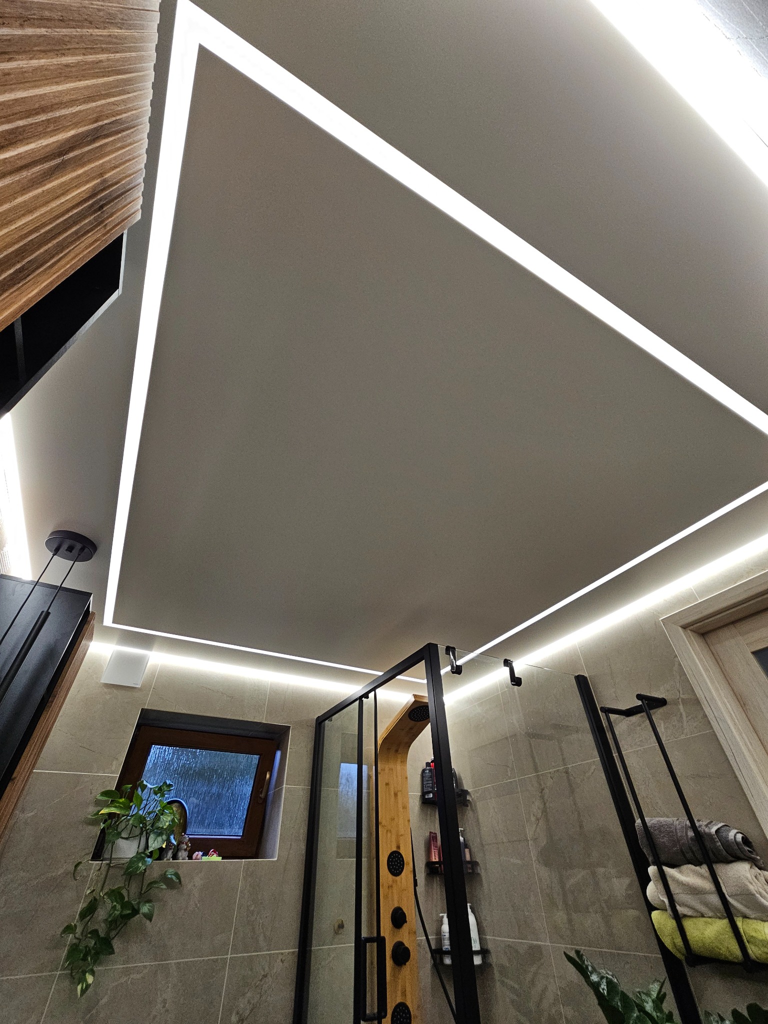 Nowoczesna łazienka z sufitem napinanym, oświetleniem LED w kształcie ramy i kabiną prysznicową. Widok z dołu podkreśla minimalistyczny design i stonowaną kolorystykę.