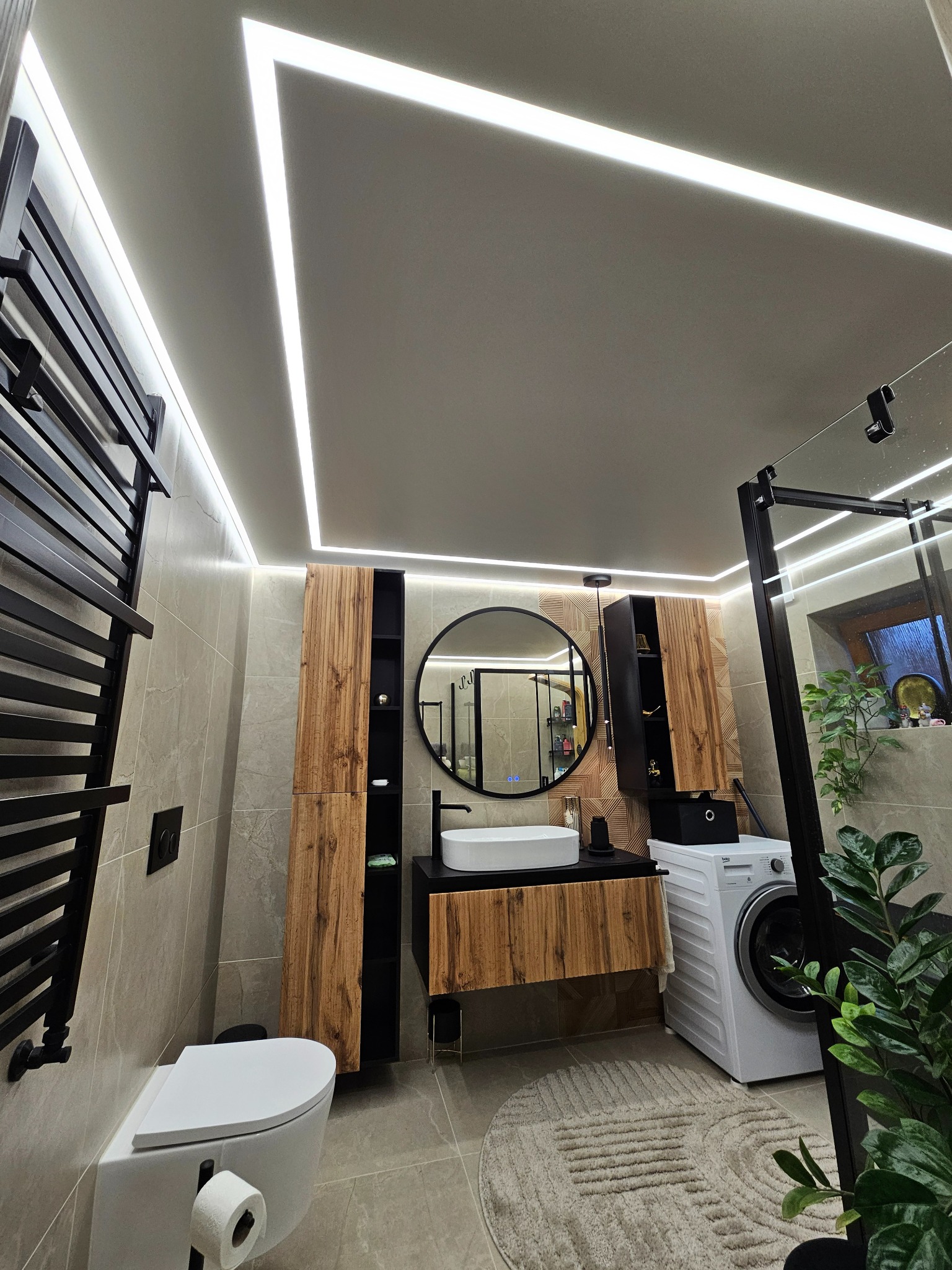 Nowoczesna łazienka z podświetlanym sufitem, drewnianymi szafkami i okrągłym lustrem. Uwagę przykuwa geometryczny wzór dywanika oraz roślinność w pomieszczeniu.