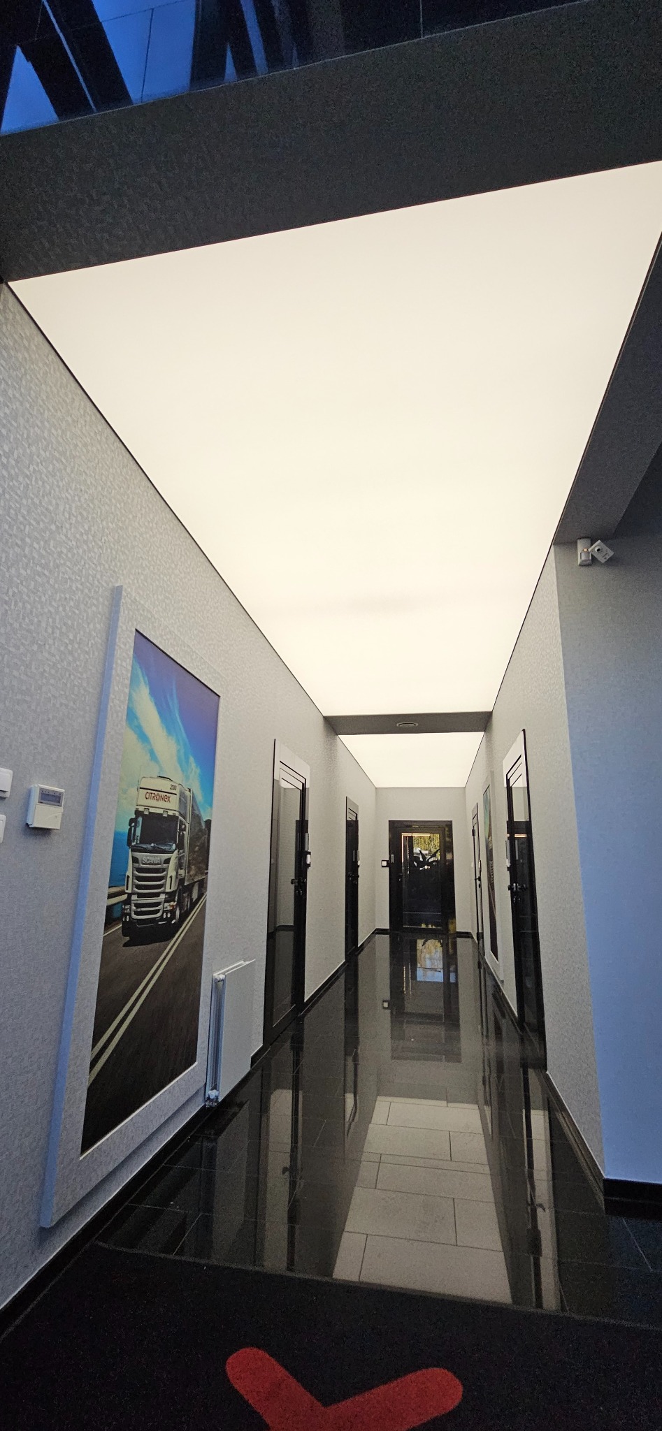 Korytarz biurowy z podświetlanym sufitem napinanym, czarne płytki na podłodze z połyskiem, grafika na ścianie. Nowoczesne wykończenie wnętrza.