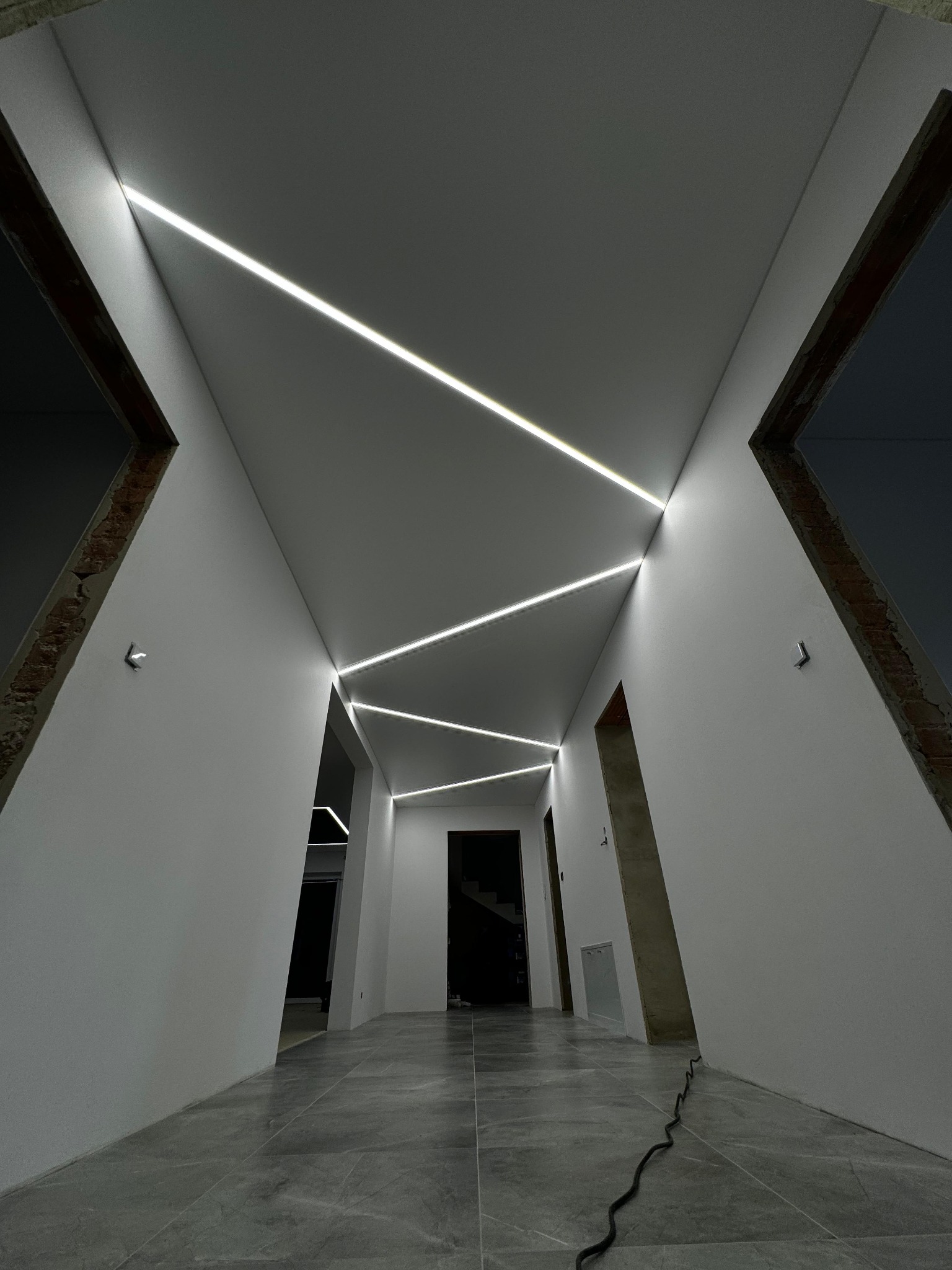 Nowoczesne oświetlenie liniowe LED w korytarzu z szarymi płytkami podłogowymi i białymi ścianami. Widok z dołu ukazuje geometryczny układ lamp.