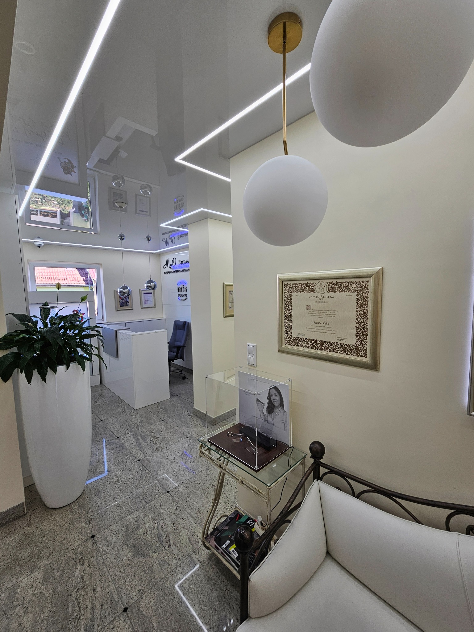 Błyszczący sufit napinany z oświetleniem LED w nowoczesnym wnętrzu. Widoczne elementy dekoracyjne, lampy wiszące i certyfikat na ścianie. Biała recepcja w tle.