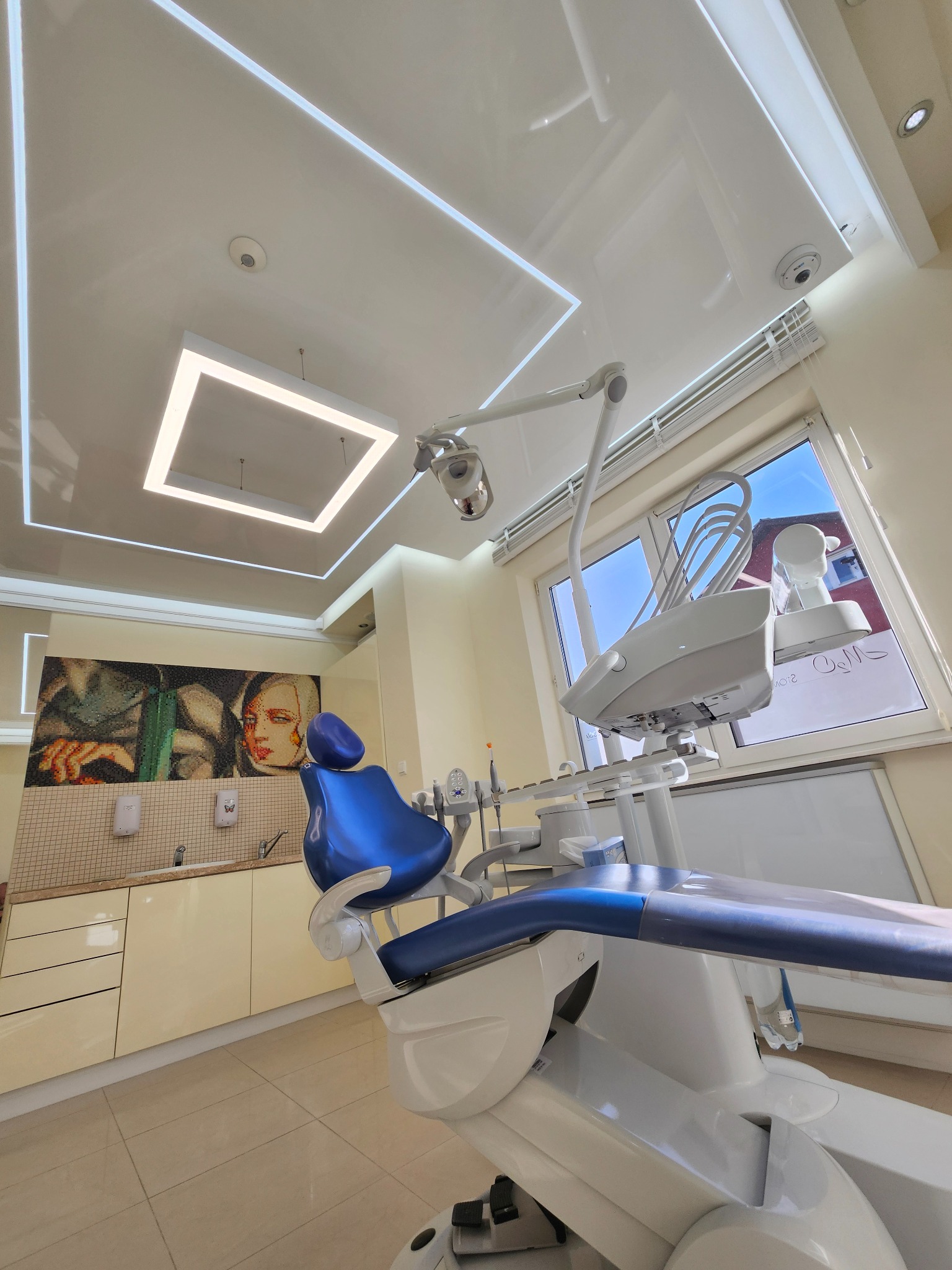 Nowoczesny gabinet dentystyczny z sufitem napinanym i oświetleniem LED, mozaiką na ścianie i fotelem dentystycznym w kolorze niebieskim. Jasne, przestronne wnętrze.