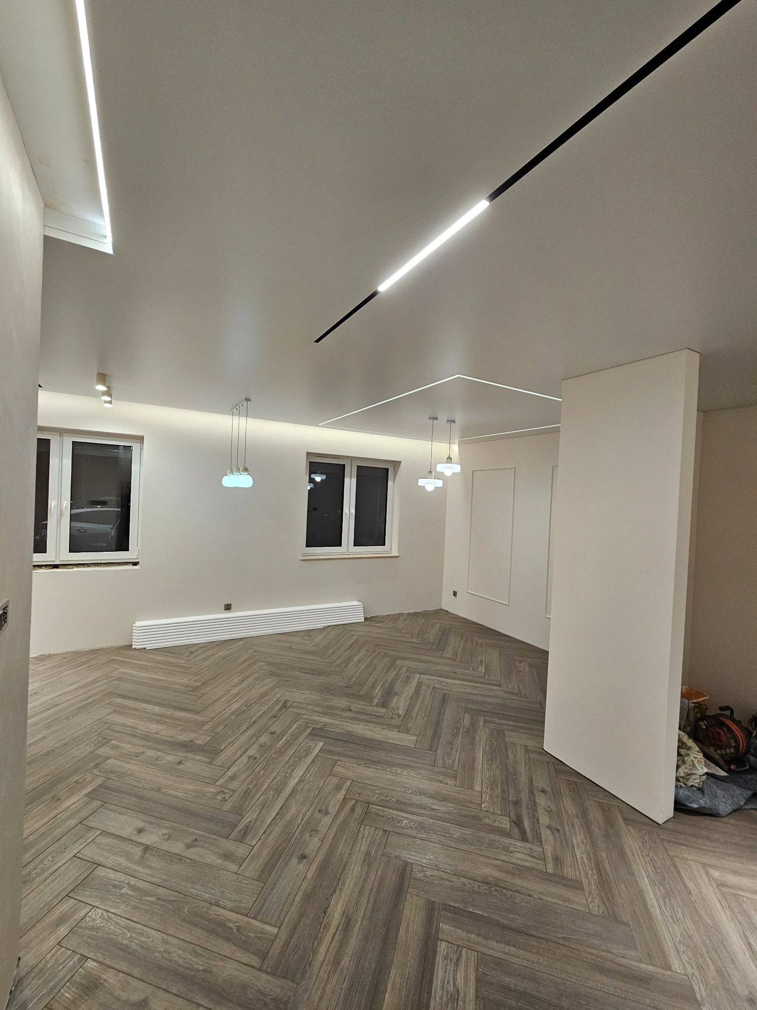 Nowoczesne wnętrze z parkietem w jodełkę, oświetleniem LED na suficie i wiszącymi lampami. Widoczne okna i grzejnik. Częściowo wykończone ściany.