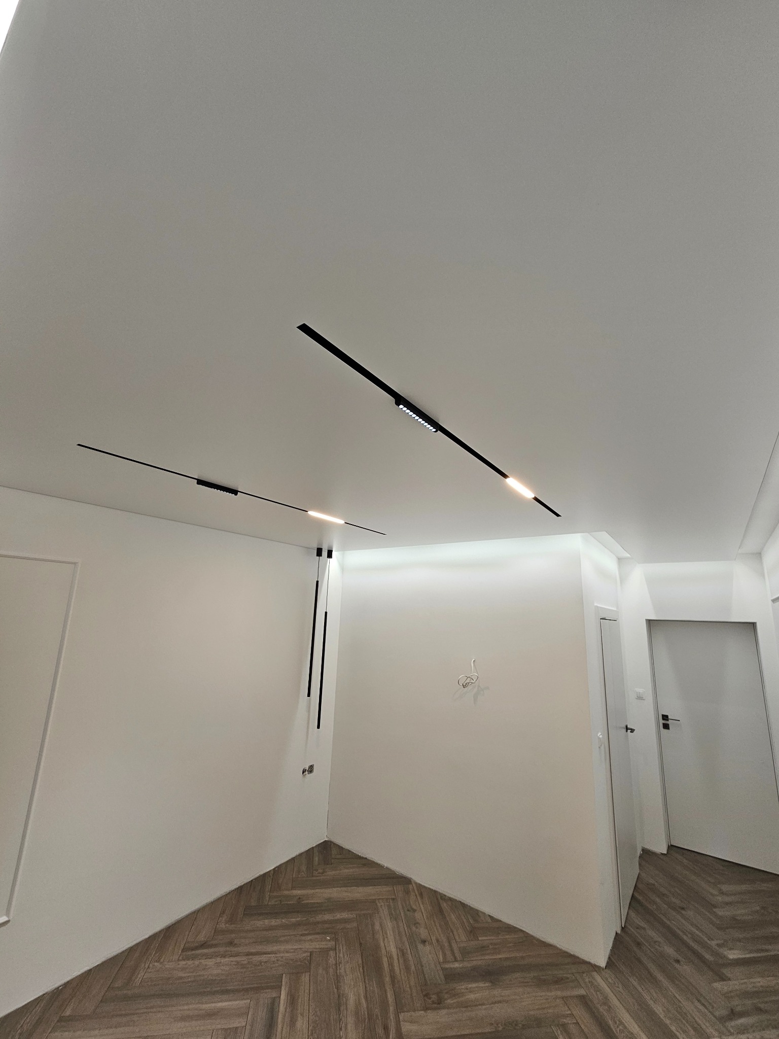 Białe wnętrze z parkietem w jodełkę i nowoczesnym oświetleniem sufitowym w postaci czarnych, liniowych lamp LED. Minimalistyczny design.