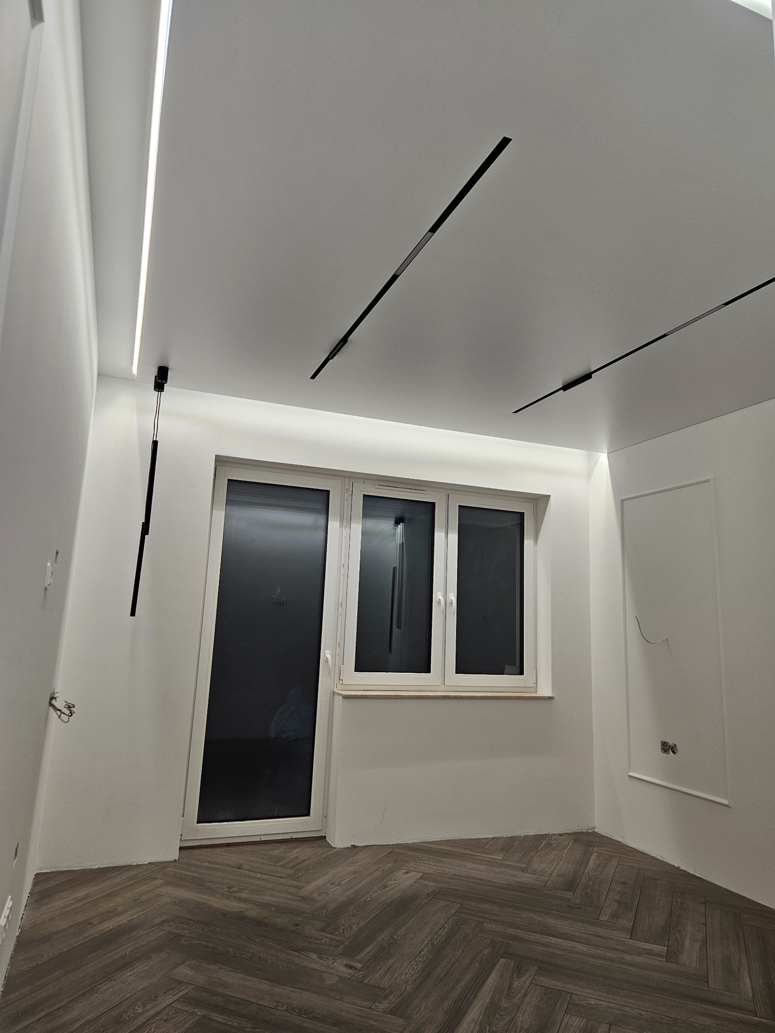 Białe wnętrze po remoncie z parkietem w jodełkę, oknem i nowoczesnym oświetleniem sufitowym: listwy LED i czarne, podłużne lampy. Widoczne gniazdka elektryczne.