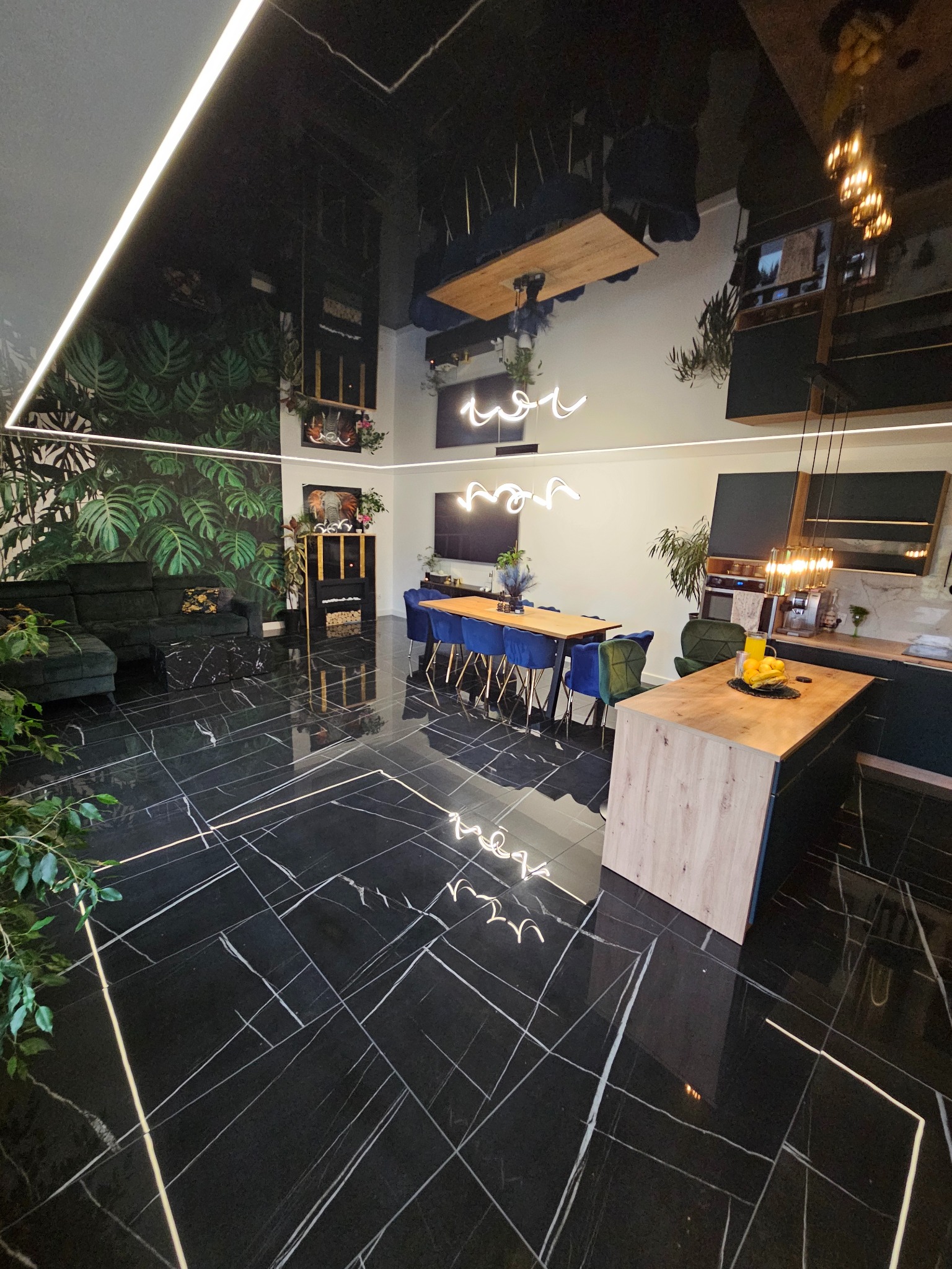 Nowoczesne wnętrze z czarną, marmurkową podłogą, sufitem lustrzanym i ścianą z motywem roślinnym. Widoczny stół z krzesłami i wyspa kuchenna z owocami.