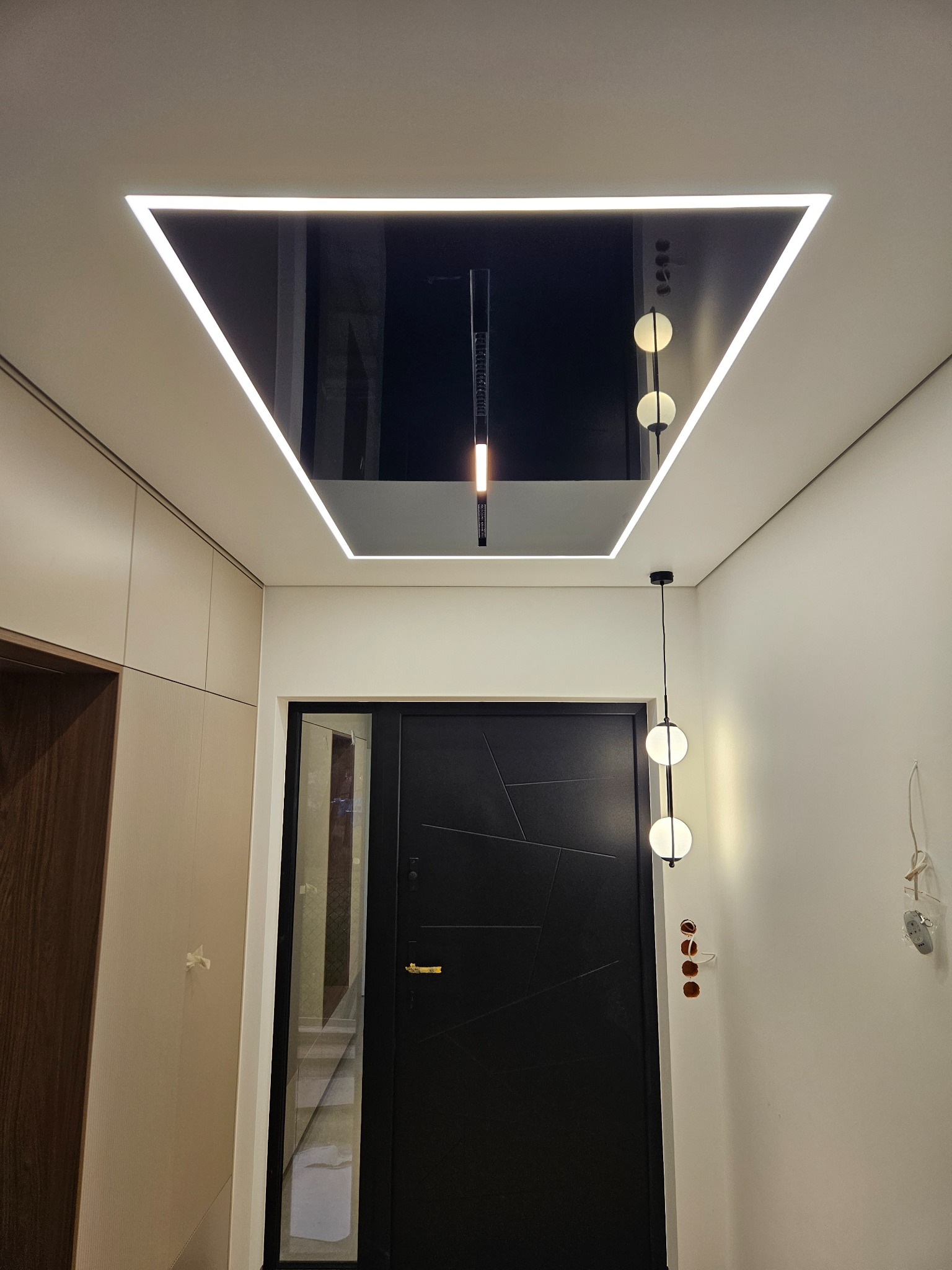Nowoczesny hol z czarnym, lustrzanym sufitem napinanym w kształcie trójkąta z oświetleniem LED. Minimalistyczny design, wisząca lampa kulista, drzwi wejściowe.