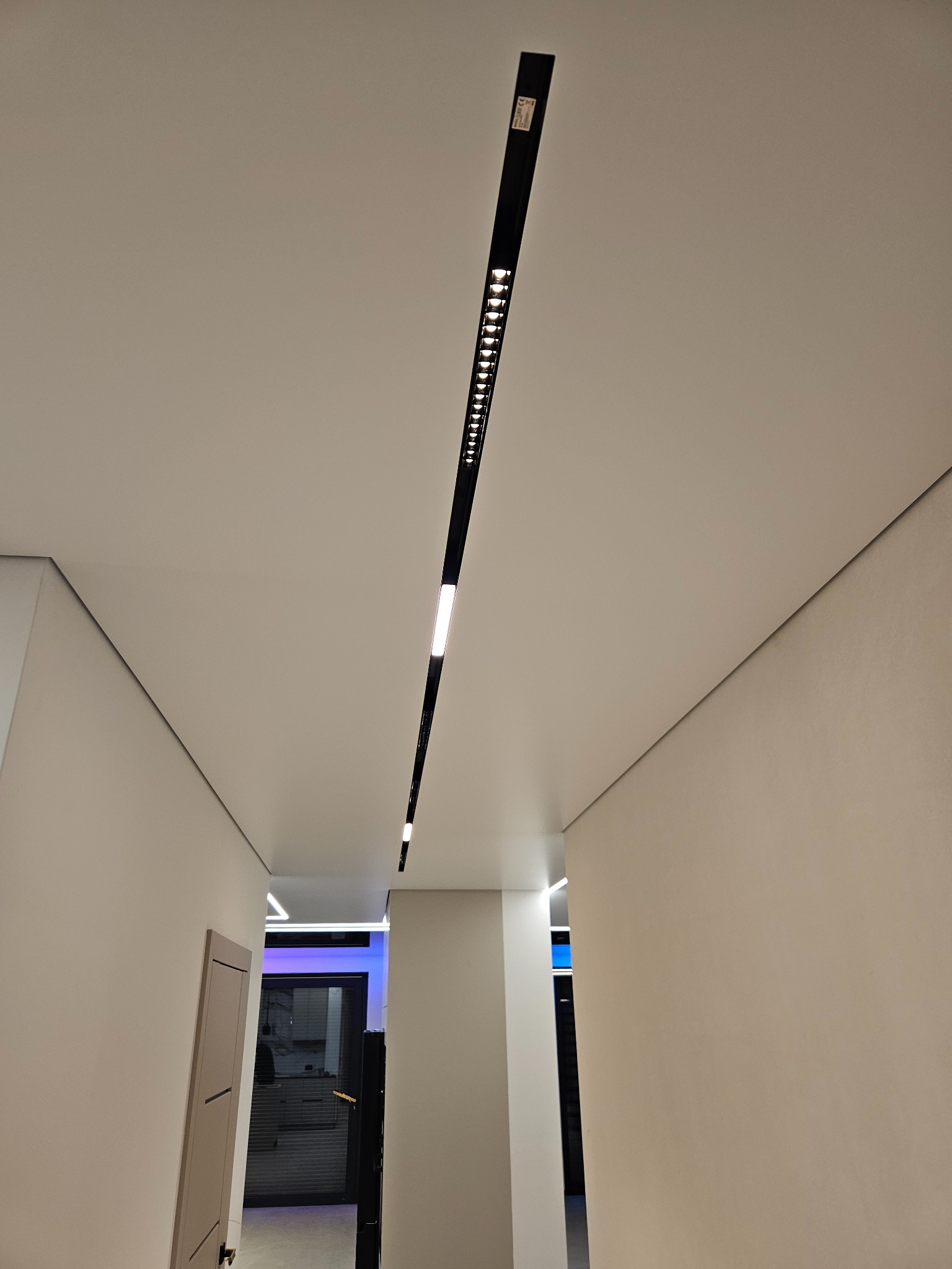 Nowoczesny korytarz z minimalistycznym oświetleniem: długa, czarna listwa LED wpuszczona w gładki, biały sufit. Ściany w jasnym odcieniu, widoczne drzwi i fragment kuchni w tle.