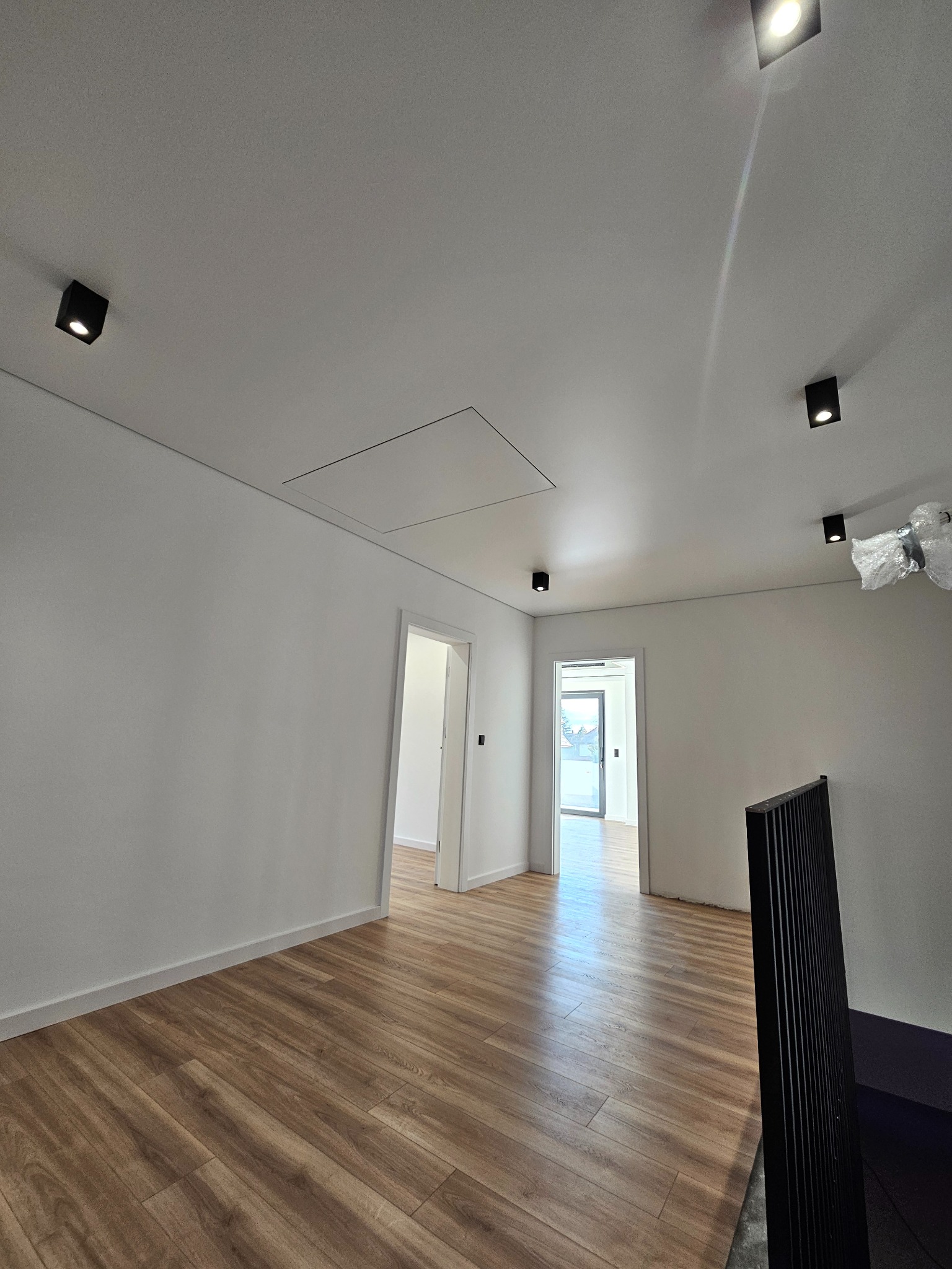 Jasne wnętrze po remoncie: drewnopodobna podłoga, białe ściany z czarnymi punktowymi lampami sufitowymi. Widoczne drzwi do kolejnych pomieszczeń.