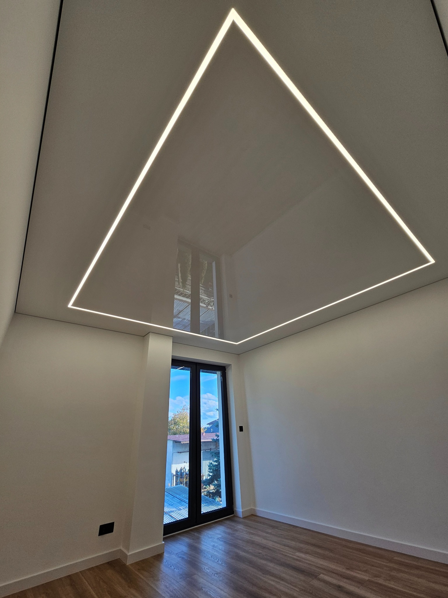 Biały, błyszczący sufit napinany z wbudowanym, kwadratowym oświetleniem LED w nowoczesnym pokoju z drewnianą podłogą i dużym oknem balkonowym.