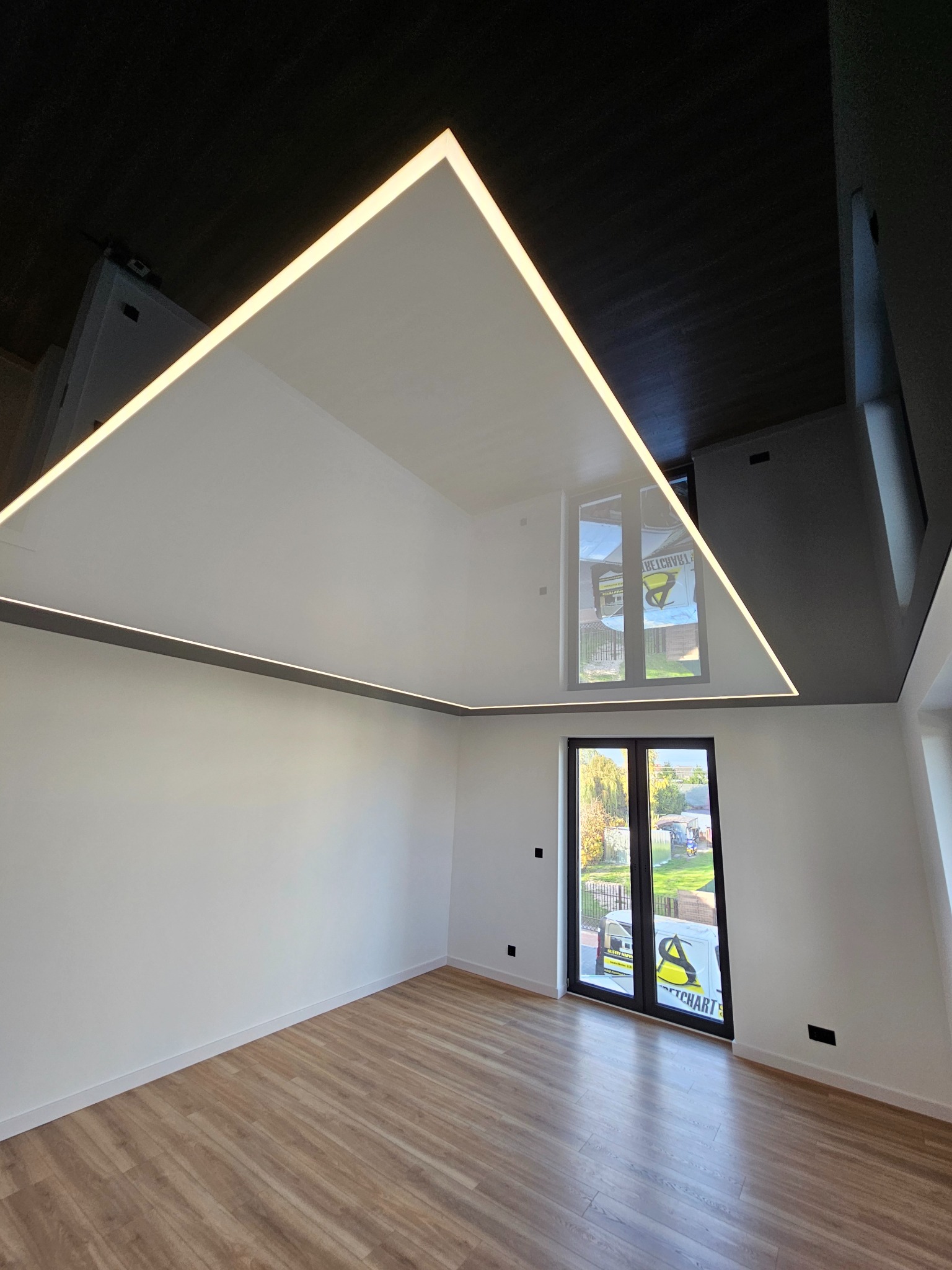 Nowoczesne wnętrze z białymi ścianami i podłogą z paneli. Uwagę przyciąga czarny sufit z podwieszanym, trójkątnym sufitem napinanym z oświetleniem LED.