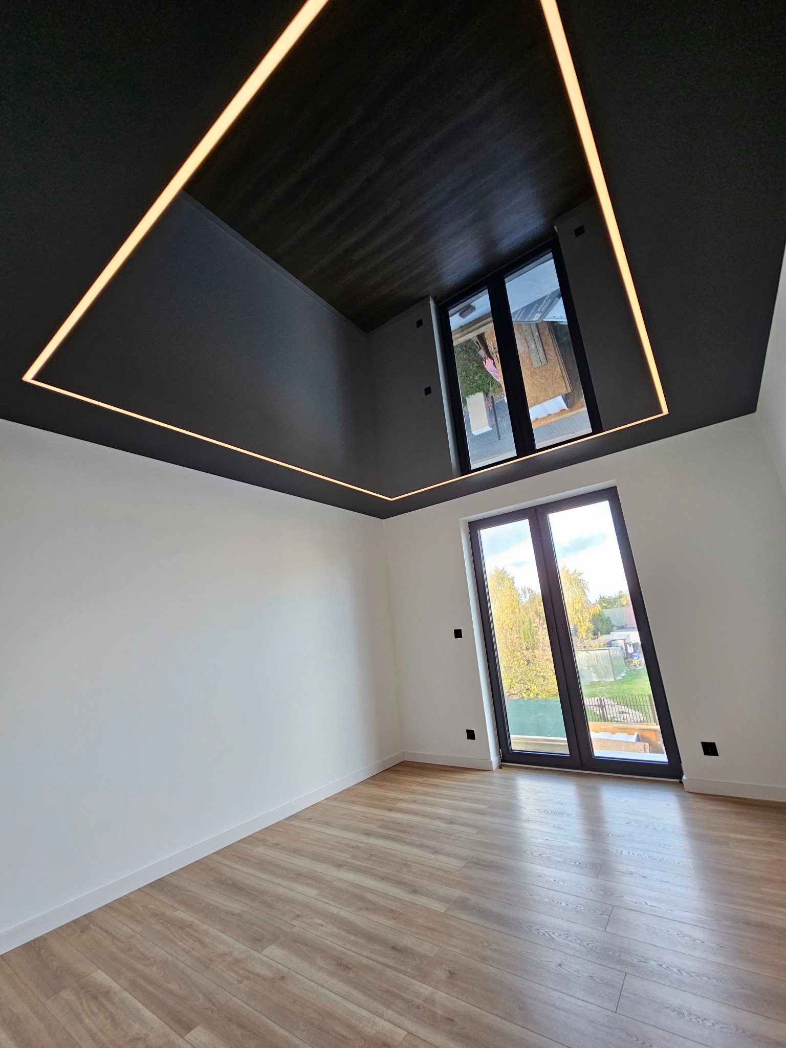 Nowoczesne wnętrze z drewnianą podłogą i czarnym sufitem podwieszanym z oświetleniem LED w kształcie kwadratu. Dwa okna wpuszczają naturalne światło.