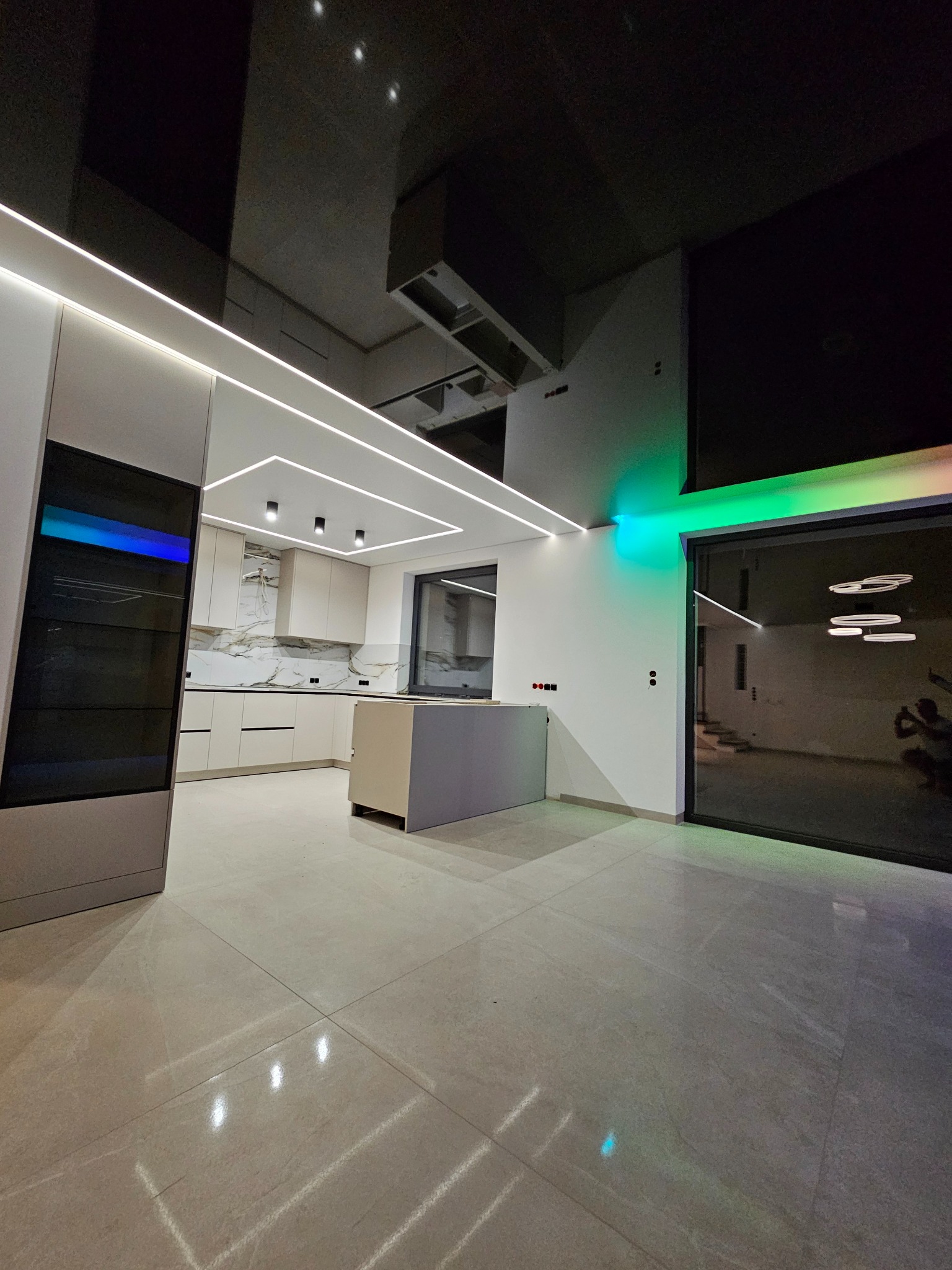 Nowoczesna kuchnia z wyspą, oświetleniem LED w suficie i pod szafkami, oraz czarnym, lustrzanym sufitem napinanym. Widoczne płytki podłogowe i duże okno.