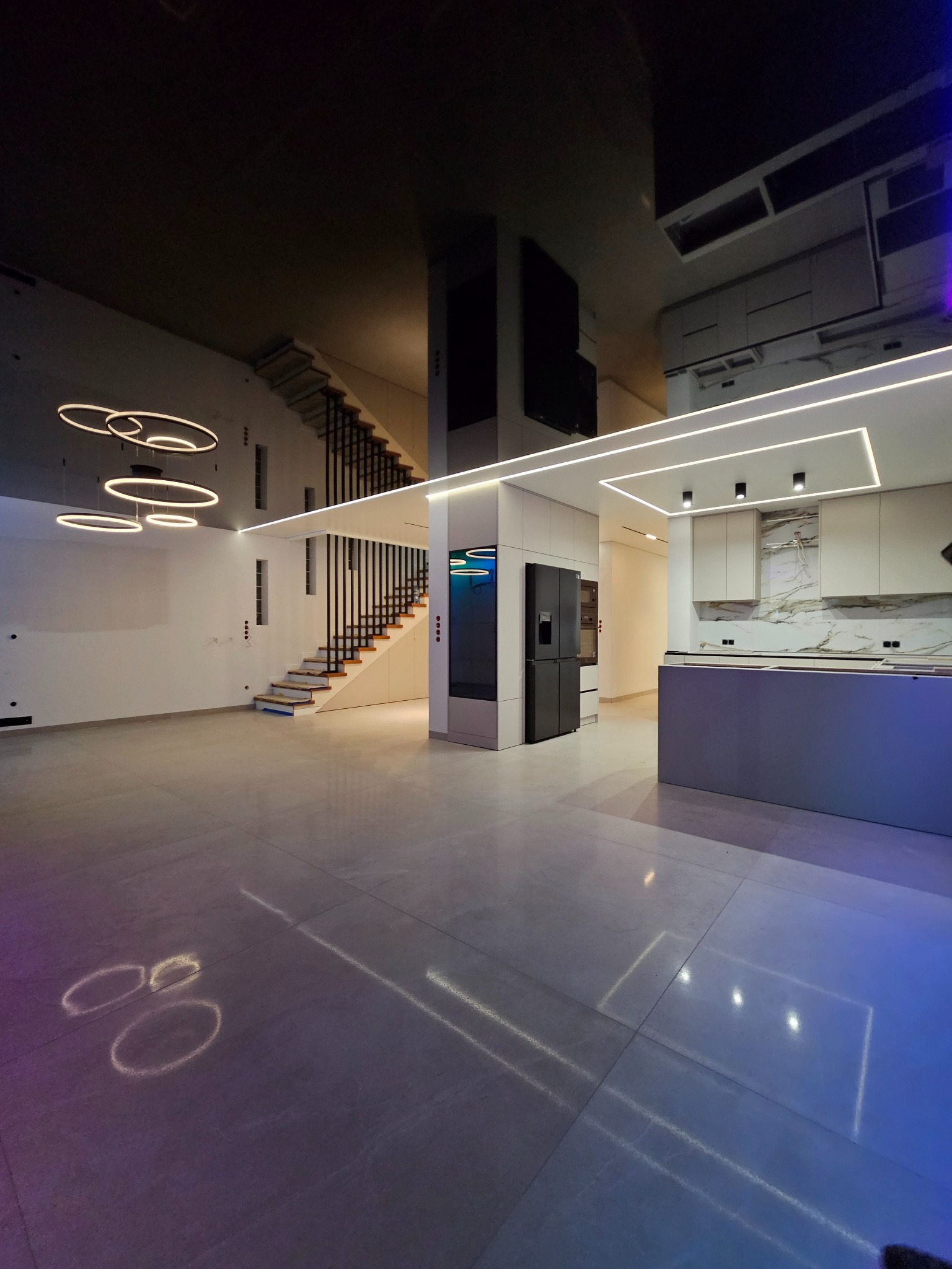 Nowoczesne wnętrze z otwartą przestrzenią: schody z drewnianymi stopniami, minimalistyczna kuchnia z wyspą i oświetleniem LED. Błyszcząca podłoga odbija światło.