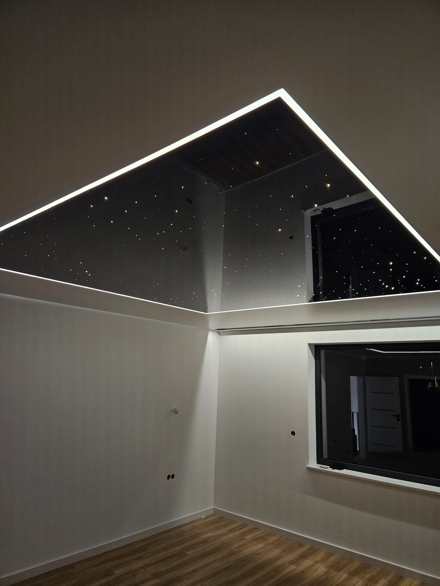 Nowoczesny pokój z czarnym sufitem napinanym imitującym rozgwieżdżone niebo, obramowanym białym oświetleniem LED. Widoczne okno i drewniana podłoga.