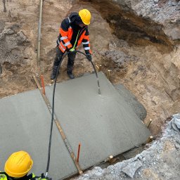 P S OSOWSCY INVESTMENTS SPÓŁKA Z OGRANICZONĄ ODPOWIEDZIALNOŚCIĄ - Wylewanie betonu w wykopie pod fundamenty z użyciem węża, pracownik w kasku i kamizelce odblaskowej. Widok z góry na plac budowy.