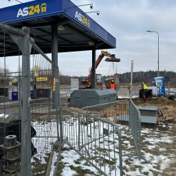 P S OSOWSCY INVESTMENTS SPÓŁKA Z OGRANICZONĄ ODPOWIEDZIALNOŚCIĄ - Montaż zbiornika na stacji paliw AS24 z użyciem koparki. Widoczne ogrodzenie placu budowy, pracownicy w kamizelkach odblaskowych i toaleta przenośna. Częściowo ośnieżony teren.