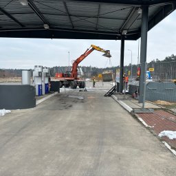 P S OSOWSCY INVESTMENTS SPÓŁKA Z OGRANICZONĄ ODPOWIEDZIALNOŚCIĄ - Czerwona koparka kołowa pod wiatą stacji paliw AS24, podnosi kontener. W tle pracownicy w kamizelkach, ogrodzenie i las. Etap prac ziemnych na stacji.