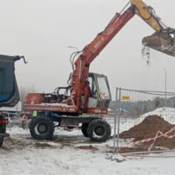 Pomarańczowa koparka MH4 wysypuje ziemię na pryzmę obok niebieskiej ciężarówki na zaśnieżonym placu budowy, ogrodzonym barierkami z tablicami ostrzegawczymi.