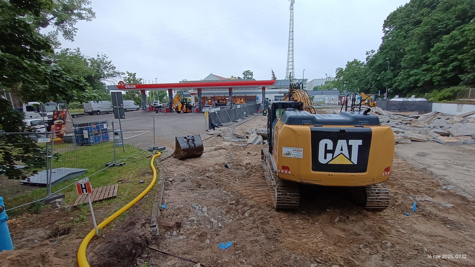 Żółta koparka CAT na placu budowy z widocznym fragmentem stacji Orlen w tle. Na ziemi leżą kamienie, a obok biegnie żółty wąż. Widok na plac budowy w dzień.