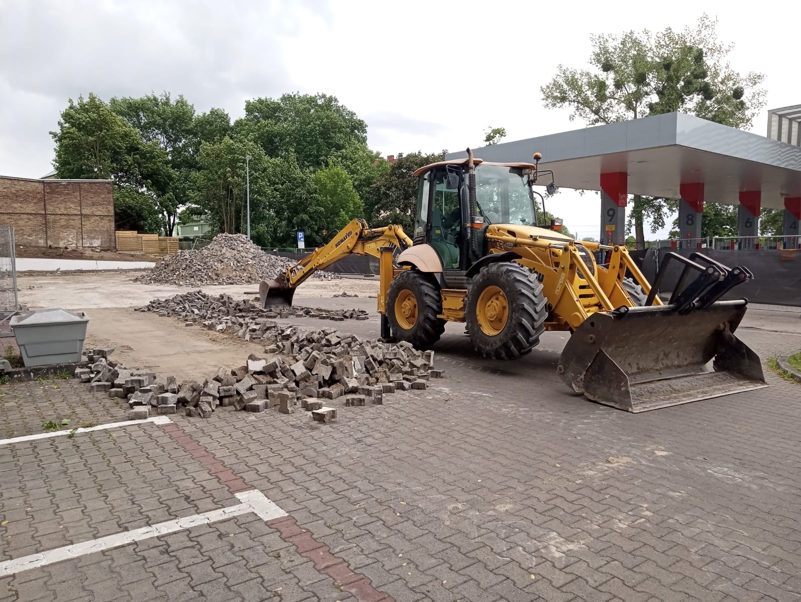 Żółta koparko-ładowarka Komatsu na placu budowy z kostką brukową, w tle stacja benzynowa i hałda gruzu. Prace ziemne na zewnątrz.