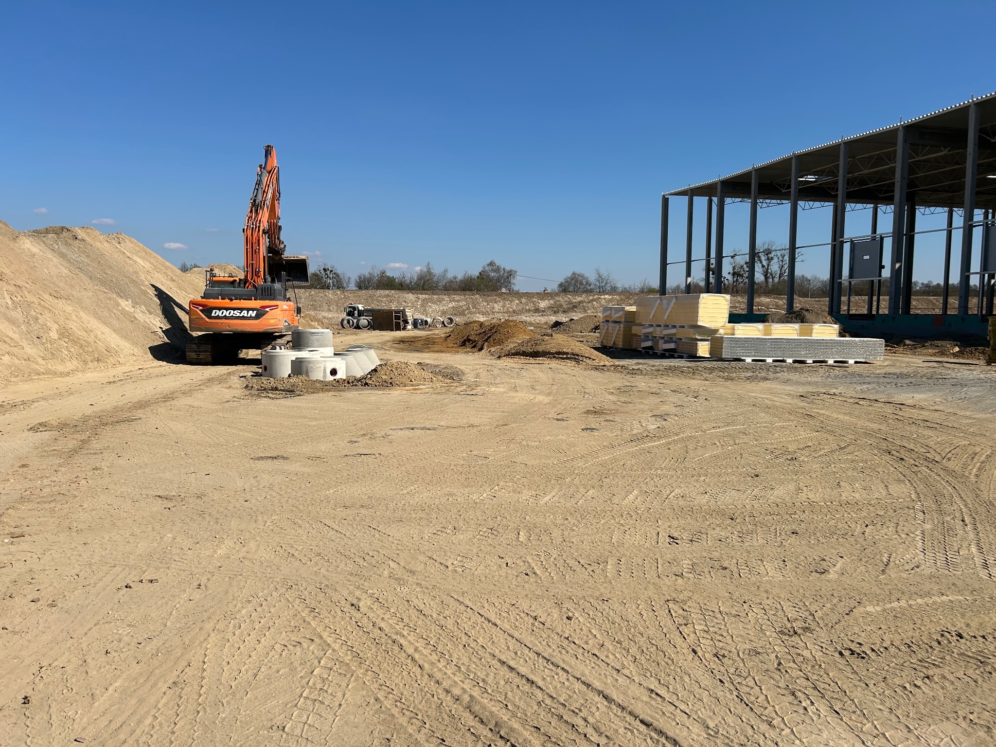 Pomarańczowa koparka Doosan na placu budowy, obok sterty ziemi i betonowych kręgów, w tle hala stalowa i błękitne niebo. Widoczne ślady opon na piasku.
