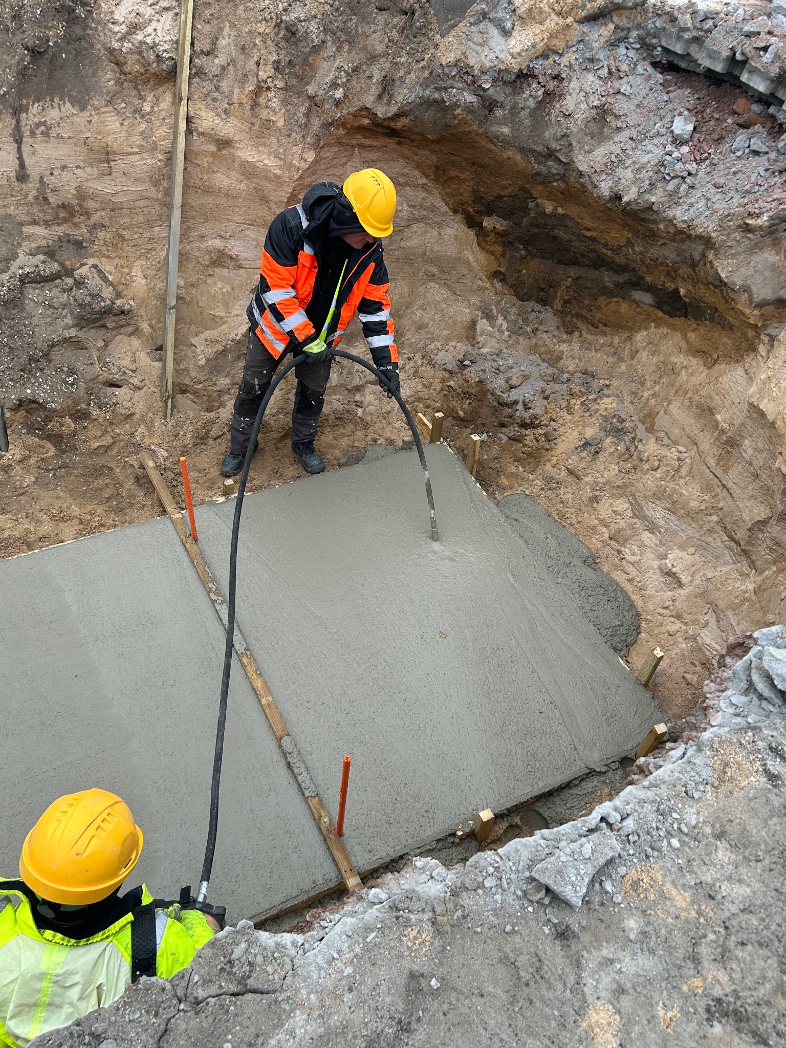 Wylewanie betonu w wykopie pod fundamenty z użyciem węża, pracownik w kasku i kamizelce odblaskowej. Widok z góry na plac budowy.