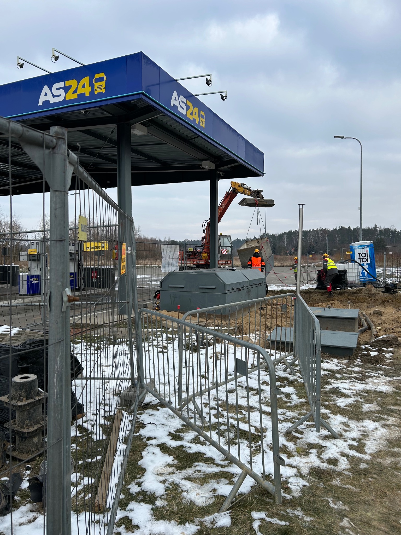 Montaż zbiornika na stacji paliw AS24 z użyciem koparki. Widoczne ogrodzenie placu budowy, pracownicy w kamizelkach odblaskowych i toaleta przenośna. Częściowo ośnieżony teren.