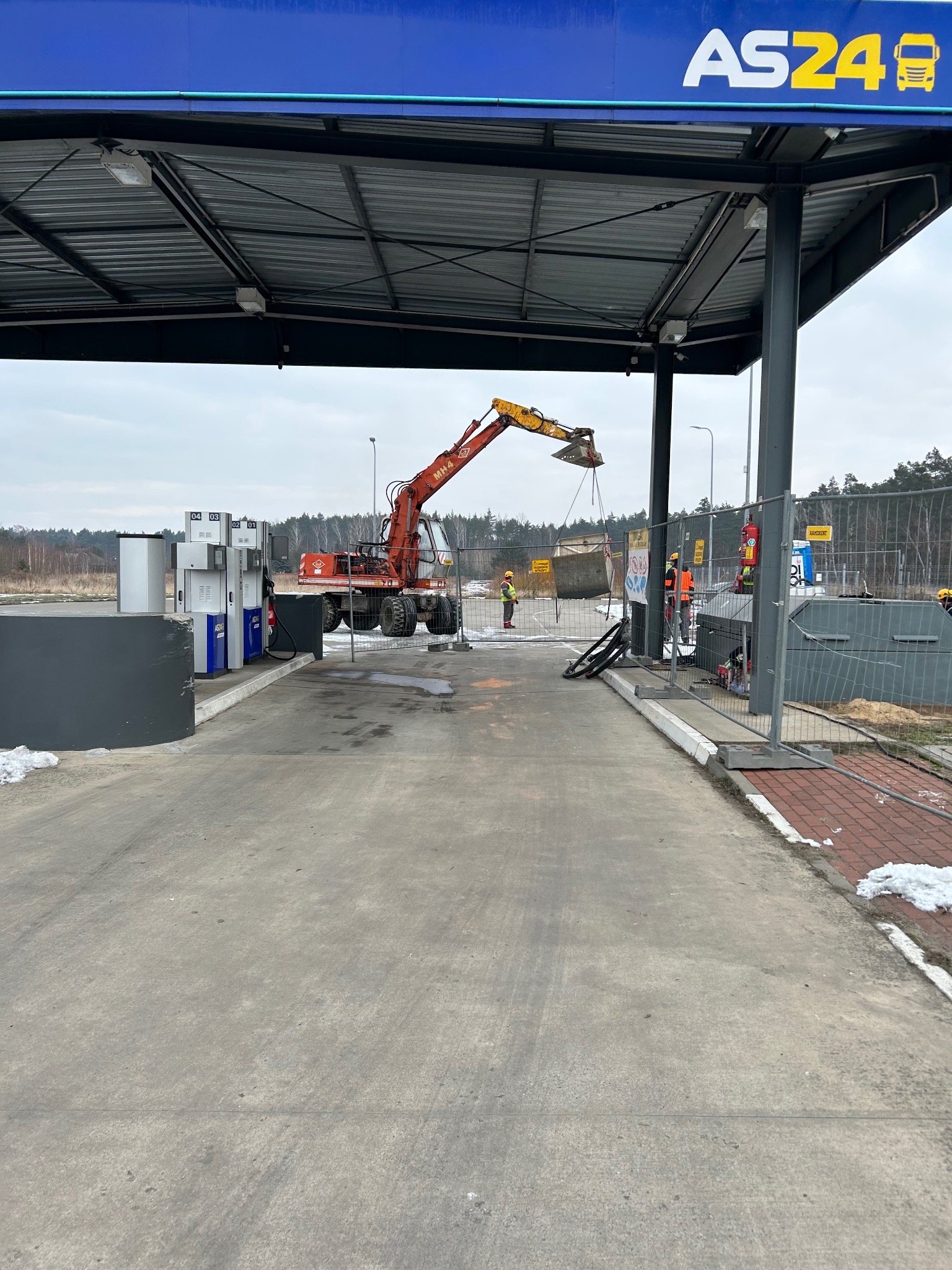 Czerwona koparka kołowa pod wiatą stacji paliw AS24, podnosi kontener. W tle pracownicy w kamizelkach, ogrodzenie i las. Etap prac ziemnych na stacji.