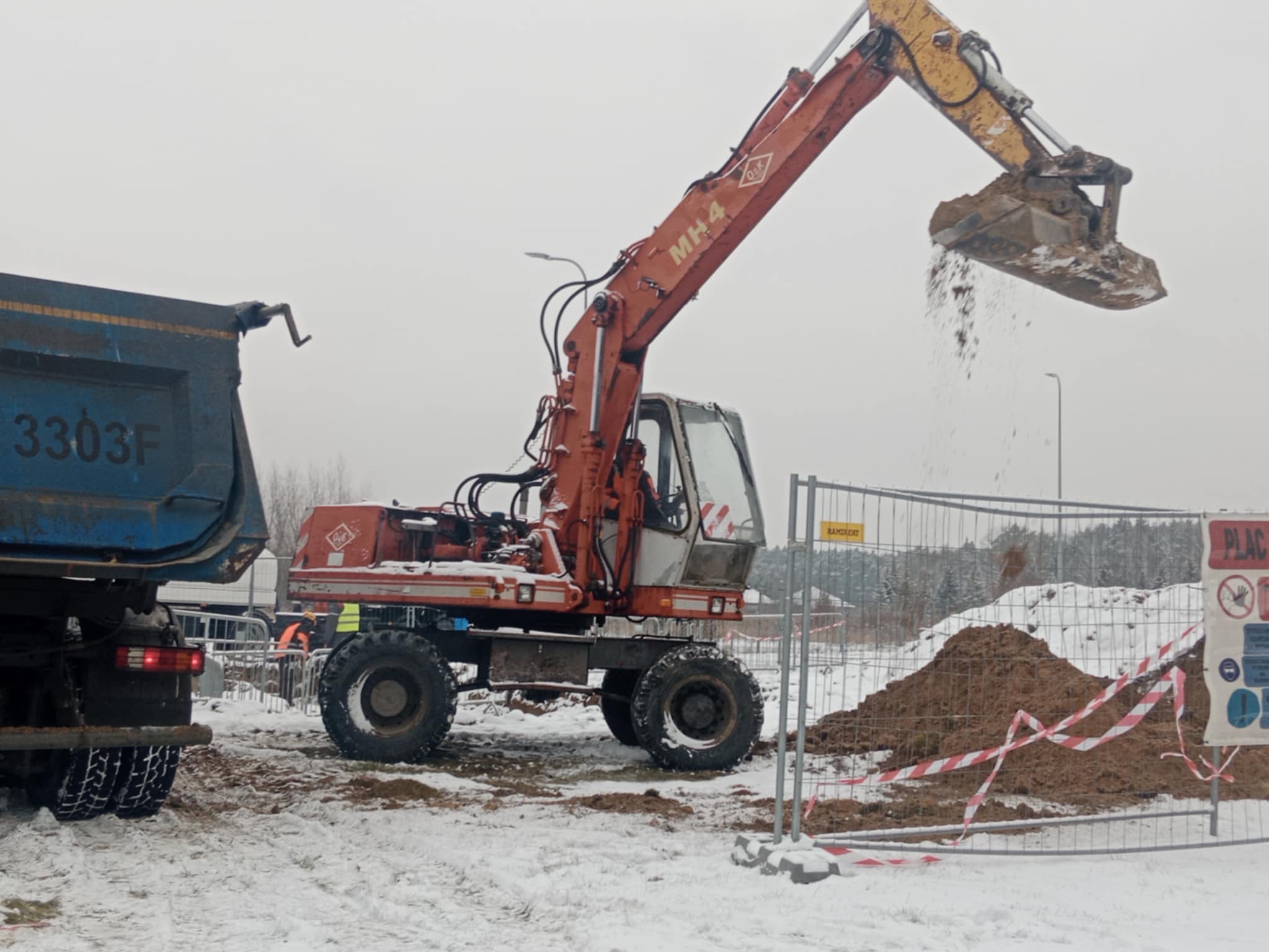 Pomarańczowa koparka MH4 wysypuje ziemię na pryzmę obok niebieskiej ciężarówki na zaśnieżonym placu budowy, ogrodzonym barierkami z tablicami ostrzegawczymi.