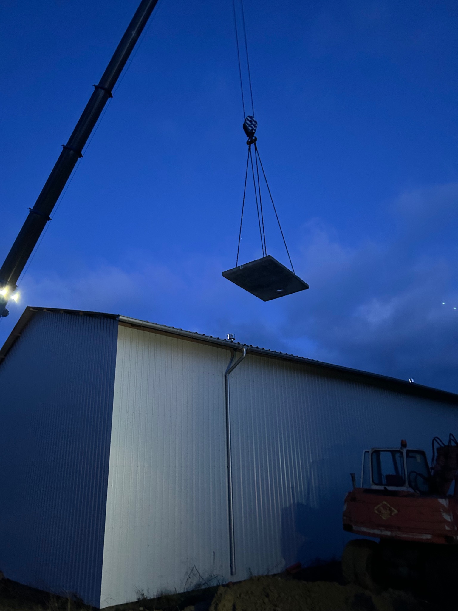 Montaż prefabrykowanego elementu betonowego dźwigiem na placu budowy przy hali w wieczornym oświetleniu. Widoczny fragment koparki.