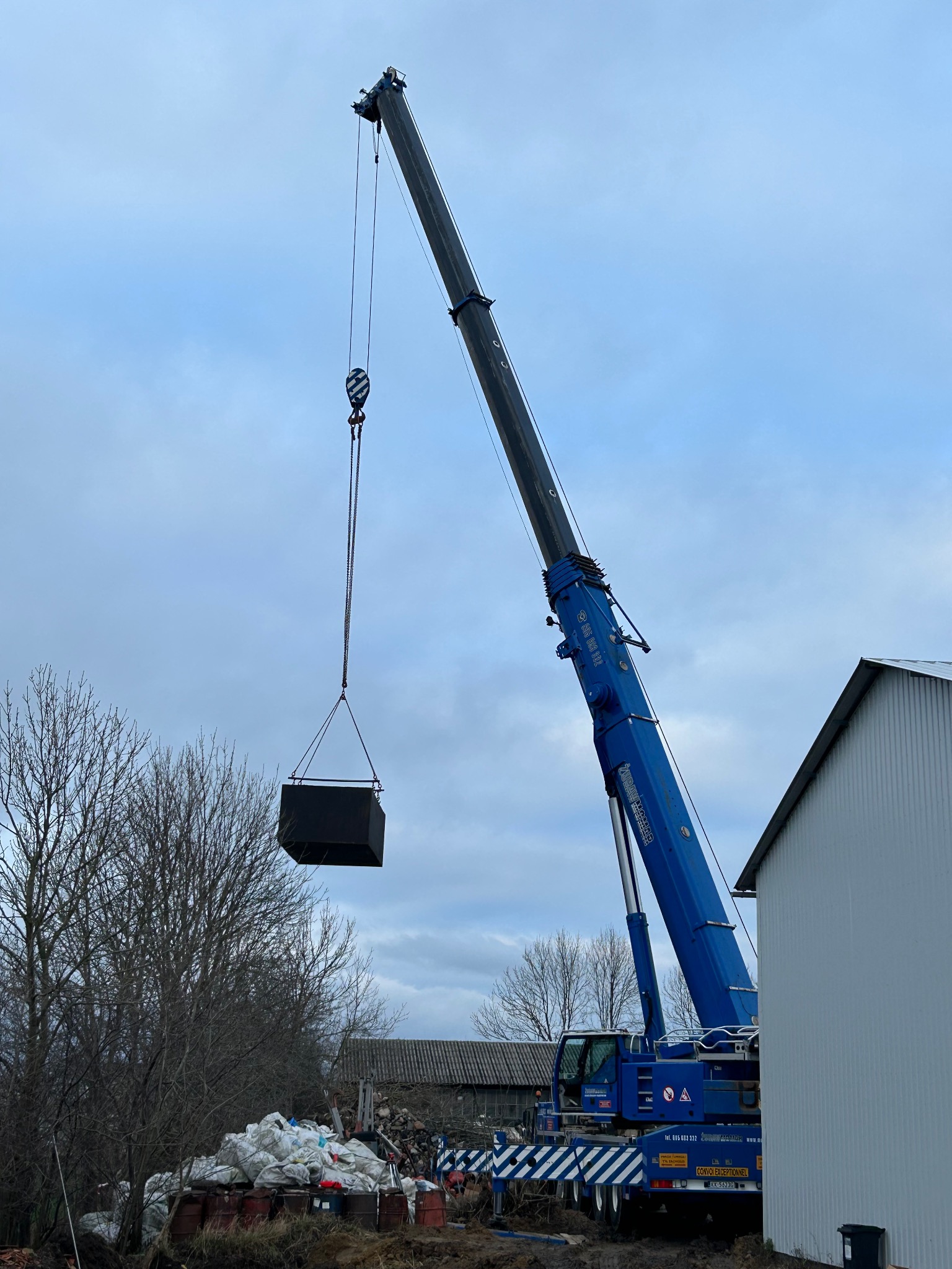 Niebieski dźwig podnosi czarny, sześcienny kontener na tle pochmurnego nieba i częściowo widocznego budynku. W tle składowane materiały budowlane.