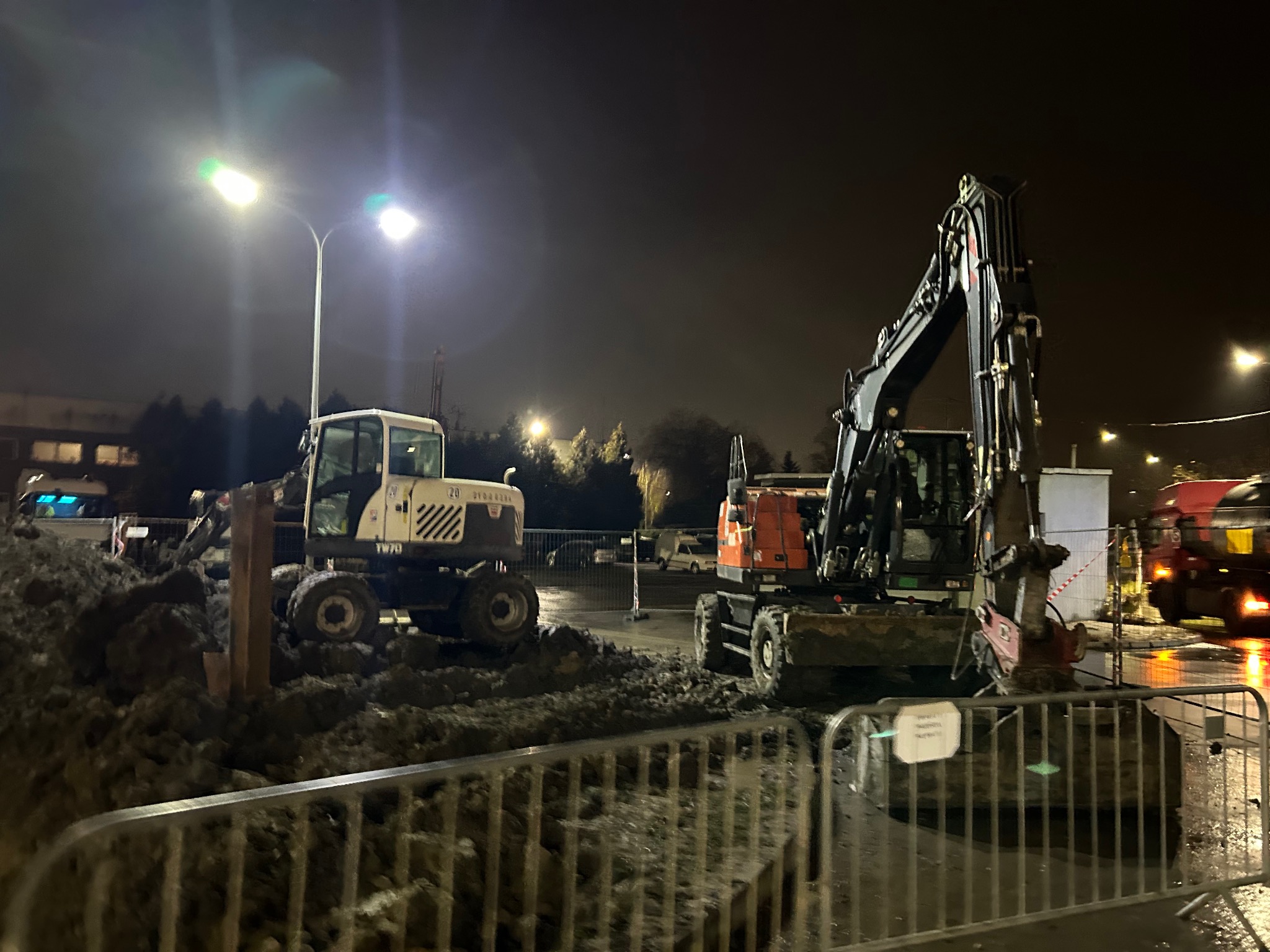 Dwa różne modele koparek kołowych na placu budowy nocą, oświetlone przez uliczne lampy. Widoczne błoto i barierki ochronne. W tle budynek i zaparkowany samochód.