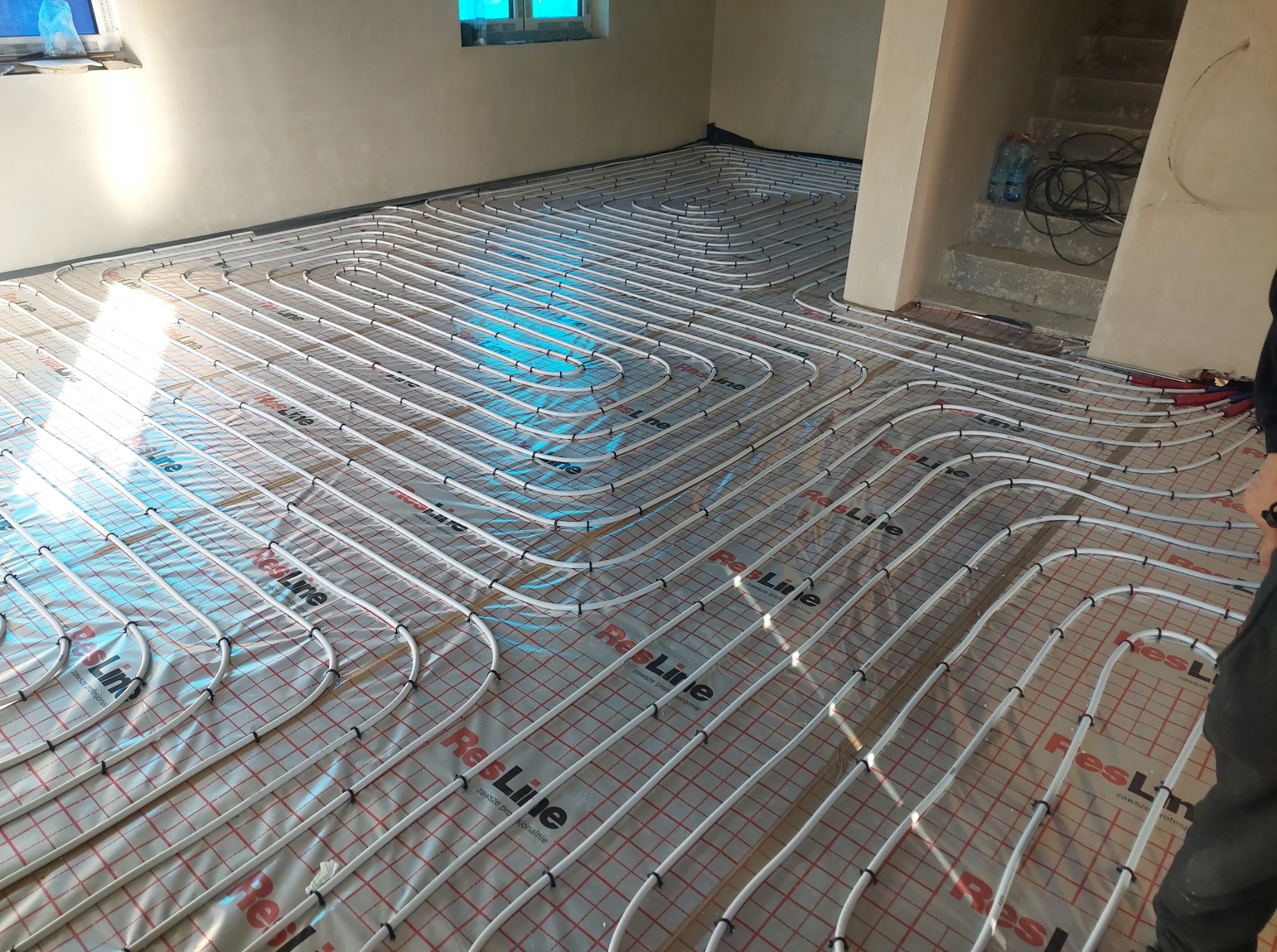 Białe rury ogrzewania podłogowego ułożone na folii z czerwoną siatką w jasnym pomieszczeniu. Widoczne fragmenty ścian i schodów. Fragment osoby po prawej stronie.