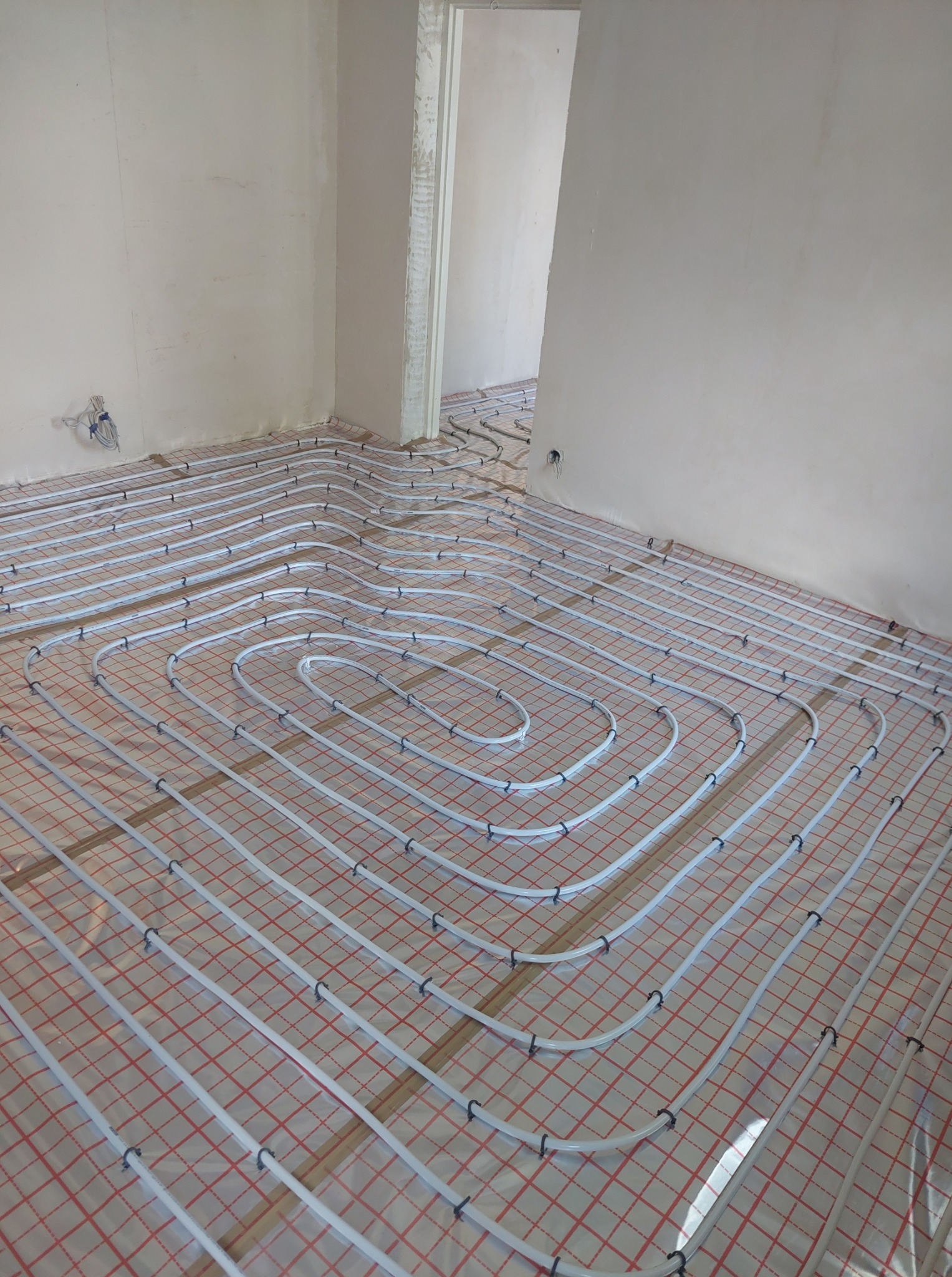 Białe rury ogrzewania podłogowego ułożone spiralnie na folii z czerwoną kratką w surowym wnętrzu. Widoczne mocowania rur i fragment ściany z instalacją elektryczną.